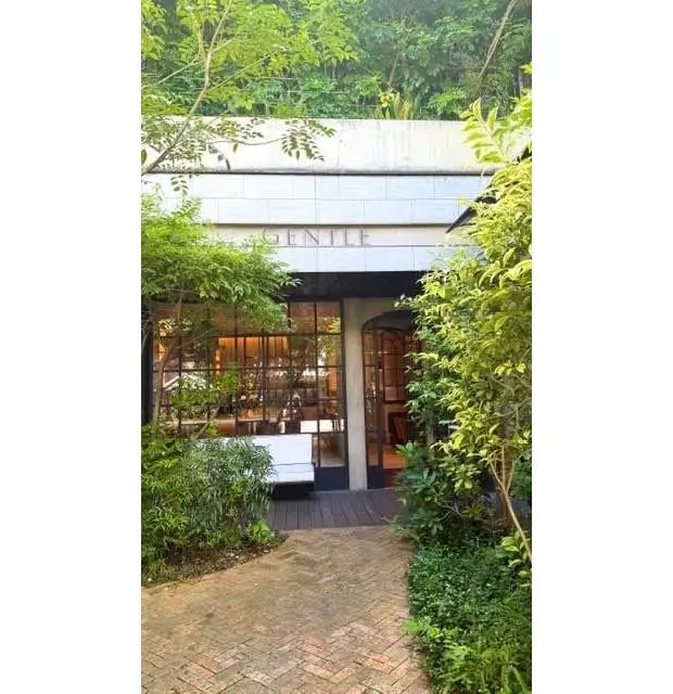 緑豊かな植栽に囲まれた、グレーの建物「Gentile」の入り口。大きなガラス窓越しに店内が見え、レンガの道が続いている。白い椅子が置かれている。