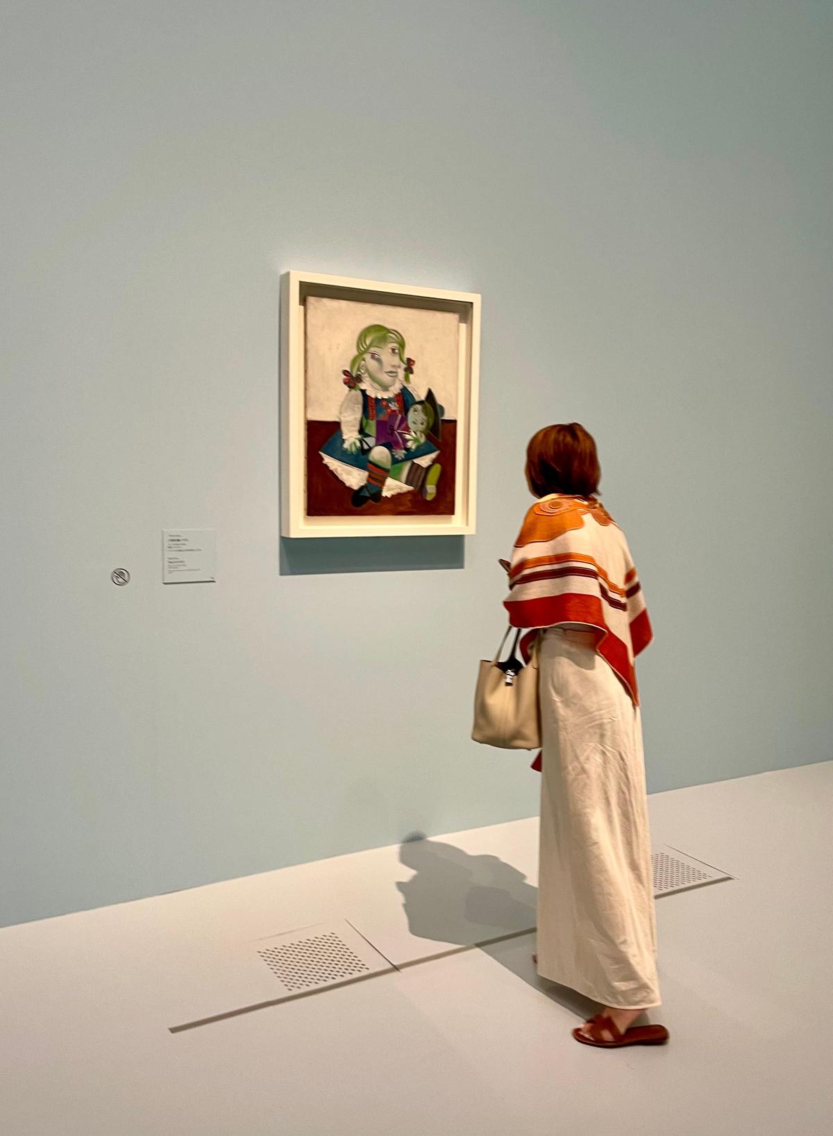 女性が白い壁に飾られた絵画を眺めている。絵画は額に入れられ、緑髪の少女と小さな人形が描かれている。女性は長袖の着物とショルダーバッグを身につけている。