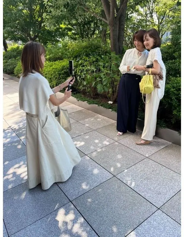 女性がモバイルカメラで、緑豊かな庭園で立ち話する二人の女性を撮影している。