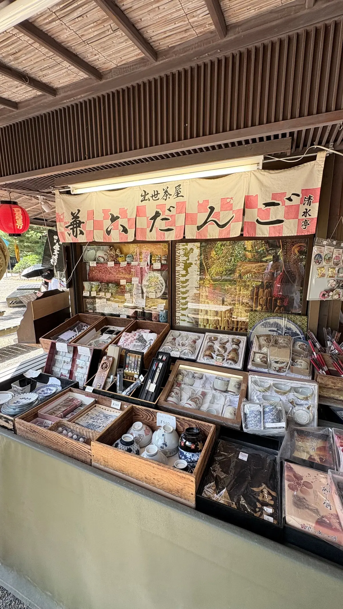 木造の屋根の下に設置された露店。木製のテーブルには、お守り、お線香、小物などが並べられ、手書きの看板が飾られている。背景には、絵画や提灯が見える。