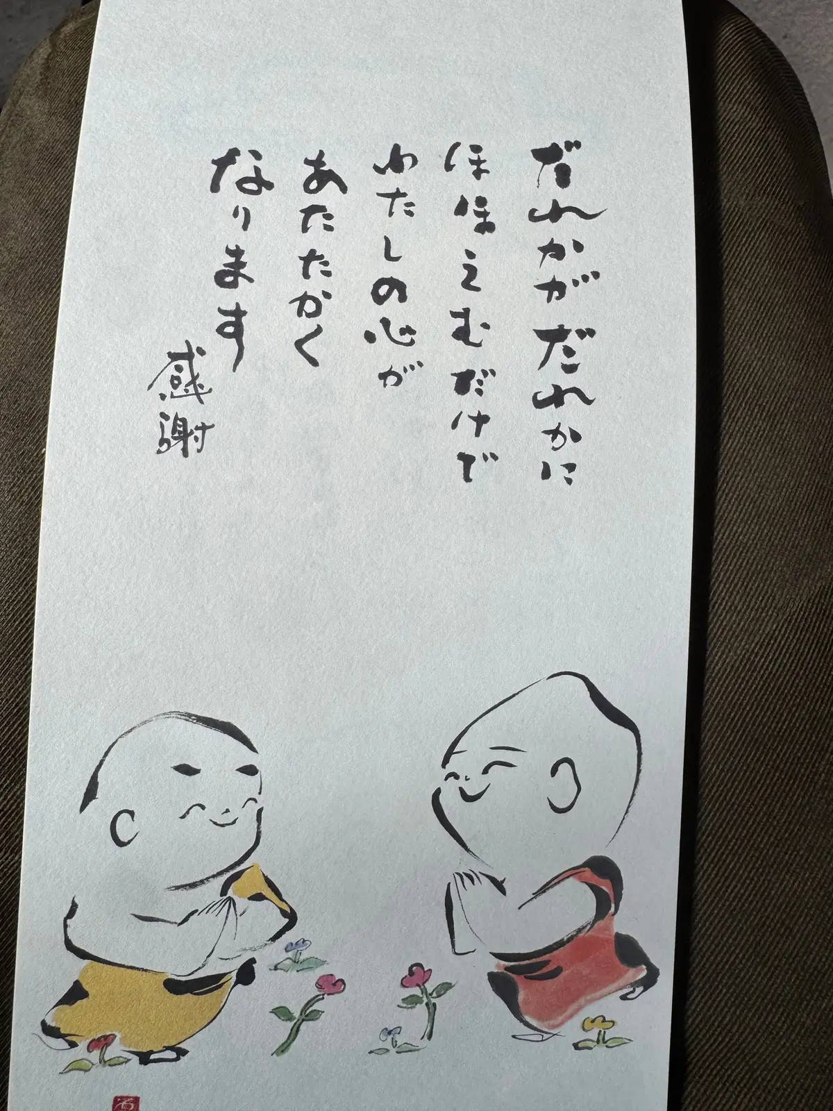 手描きの漫画風イラストと日本語の文字が書かれた紙。左側に丸顔の男性が黄色い服を着て微笑み、右側に丸顔の女性が赤いスカートを着て座っている。両者の間には小さな花が描かれている。