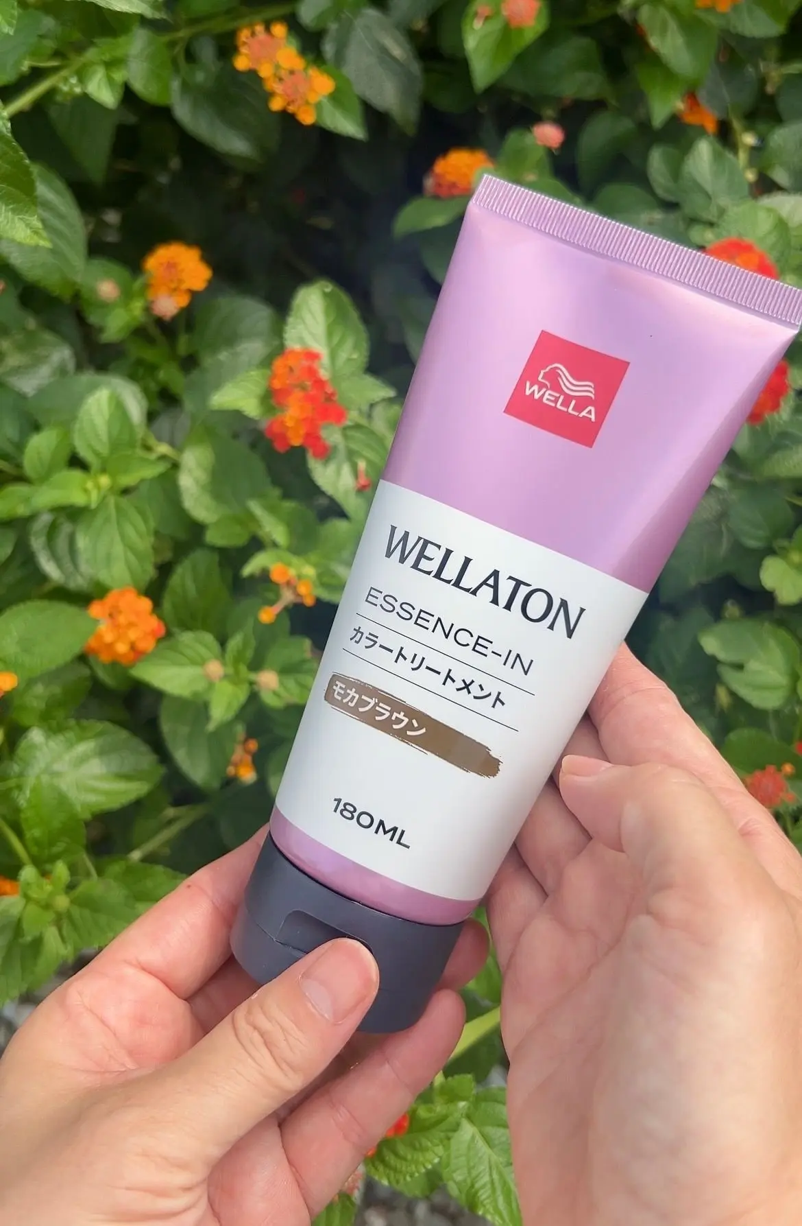 手で握られた、ウェラ(Wella)の「ウェラトーン エッセンス・イン モカブラウン」のヘアカラー剤。ピンクと紫のチューブで、180ml。背景にはオレンジ色の花が咲く緑の葉。