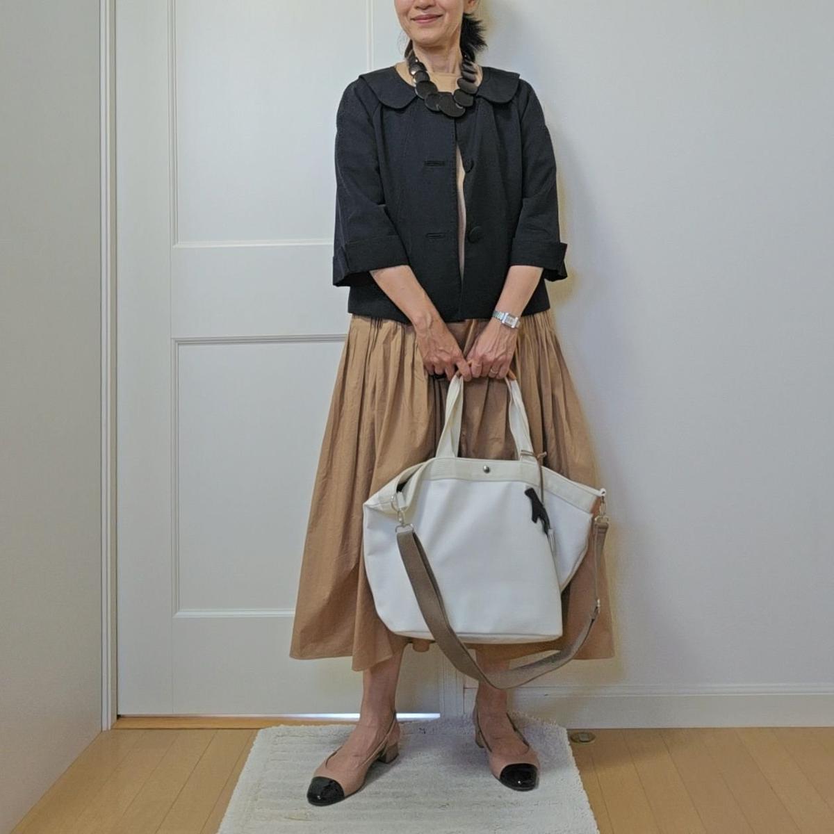 女性が黒の襟付きシャツとベージュのロングスカートを着て、笑顔で白のトートバッグを手に持っている。足元はベージュのヒールで、白いカーペットの上に立っている。