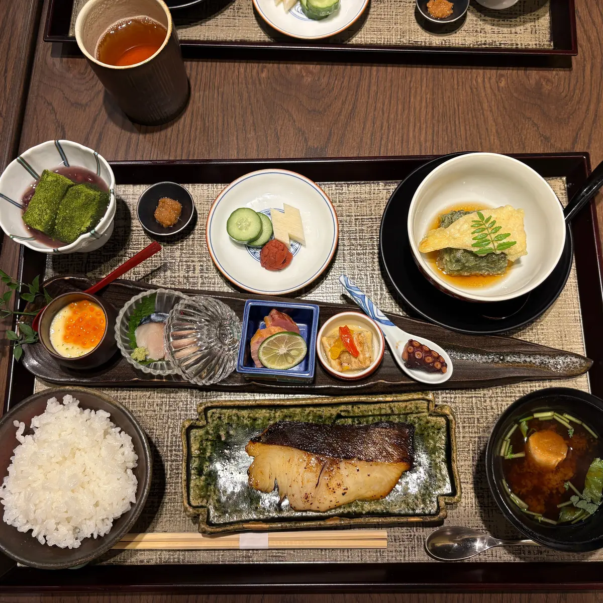 木製トレイに多様な料理が並ぶ。白米、焼き魚、味噌汁、卵焼き、刺身、ワサビ、海老、野菜の盛り合わせなど。
