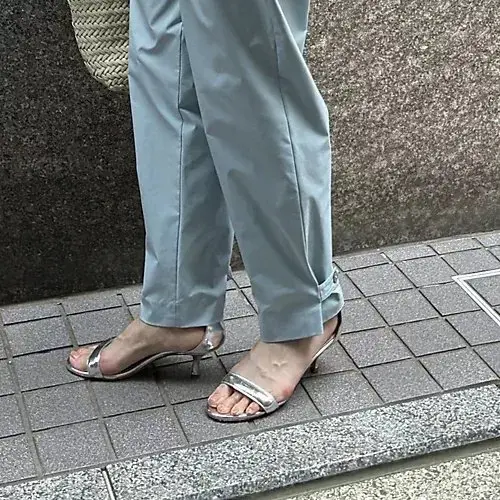 ライトブルーのパンツを履いた女性の足元。シルバーの細いストラップサンダルを履いている。足の指先がわずかに見える。背景にはコンクリートの壁とタイル張りの道。