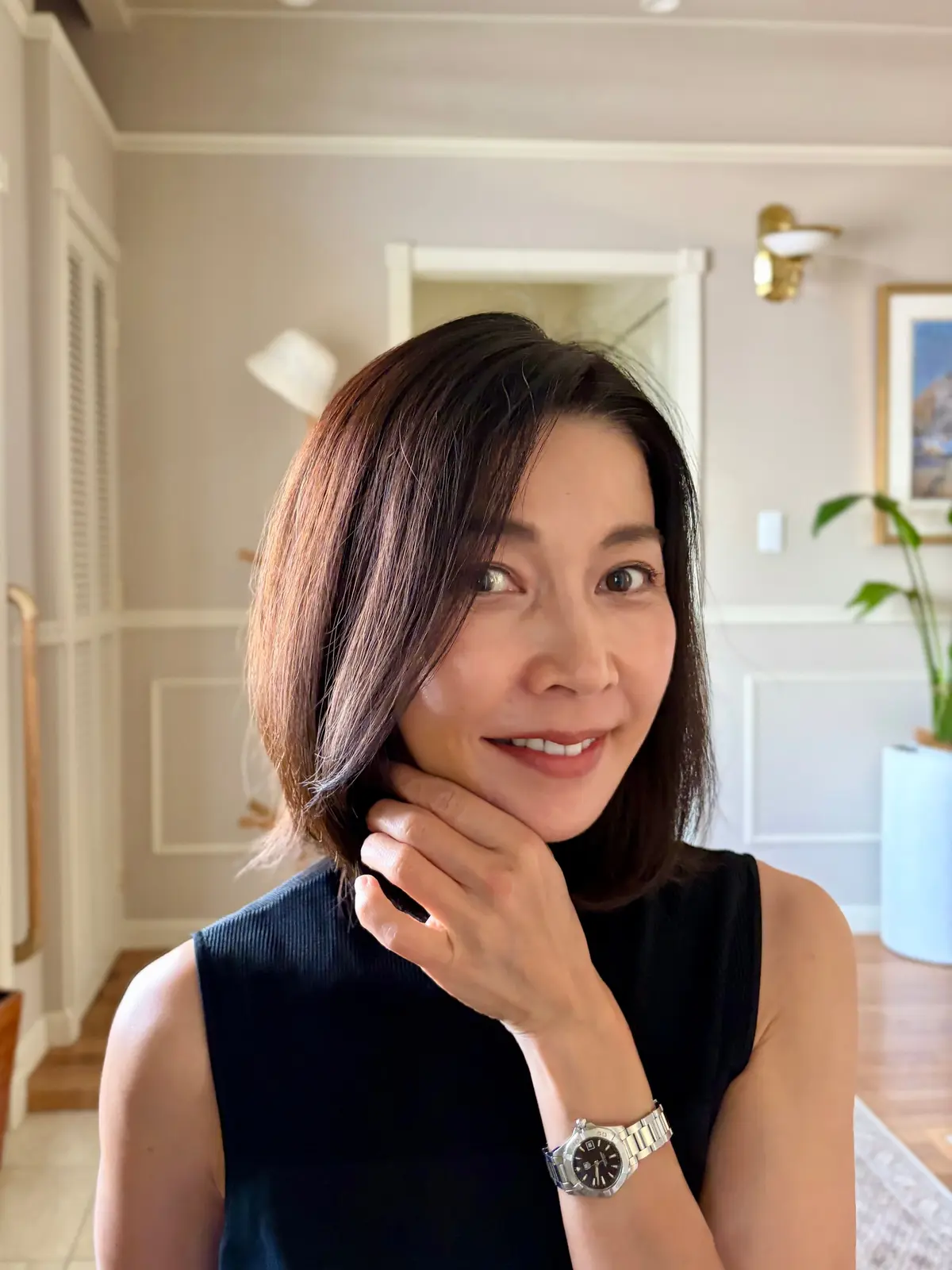 黒いタンクトップを着たアジア系の女性が、微笑みながらこちらを見ている。茶色のボブヘアで、左手を顎に当て、銀色の腕時計をしている。背景には白いドアと観葉植物が見える。