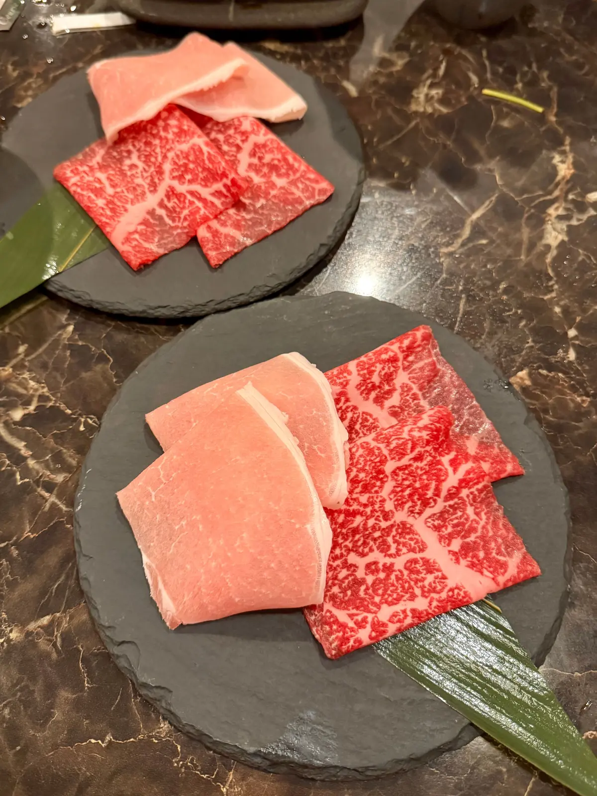 黒い石板に盛り付けられた、赤身と脂身が美しい和牛の薄切り肉。2枚の石板にそれぞれ肉が並び、緑色の薬味が添えられている。
