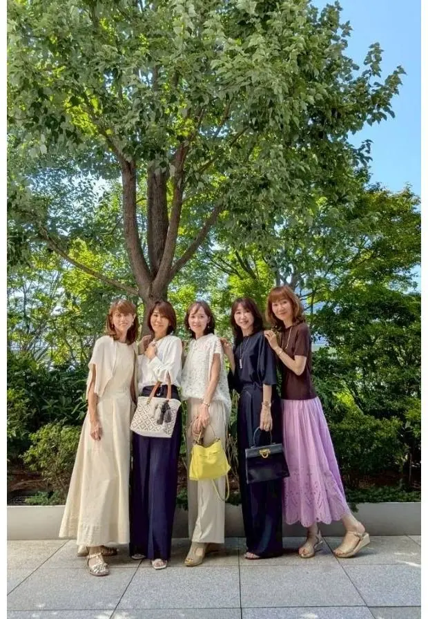 5人の女性が並んで立っており、背景には緑豊かな木が広がっている。女性たちはそれぞれ異なる服装で、左からベージュのワンピース、ネイビーのスーツ、ライトグリーンのワンピース、黒のロングスカート、ライラックのスカートを着用している。