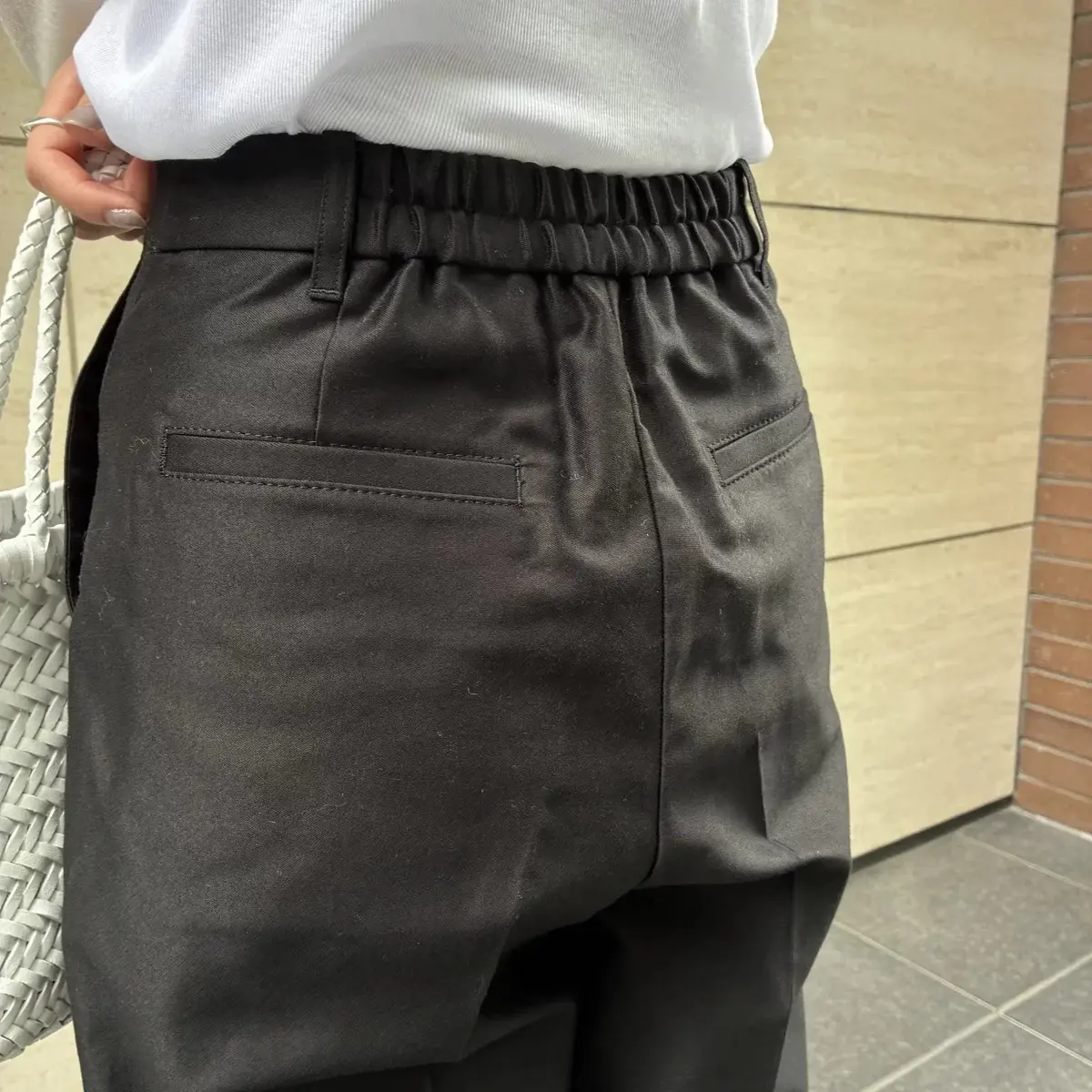 黒いサテン素材のパンツ。ウエストはゴムとドローコードで調整可能。バックポケットが2つ付いており、白いトップスと持ち合わせが映り込んでいる。