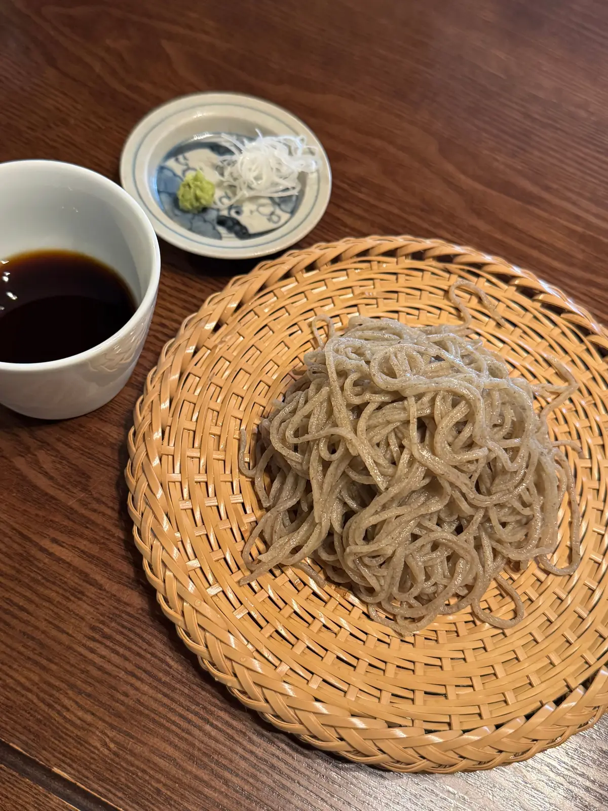 茶色い蕎麦が、丸い麦わら製の皿に盛り付けられている。そばつゆが白い器に入り、小皿には山椒とわさびが添えられている。木製のテーブルの上で撮影された料理の写真。