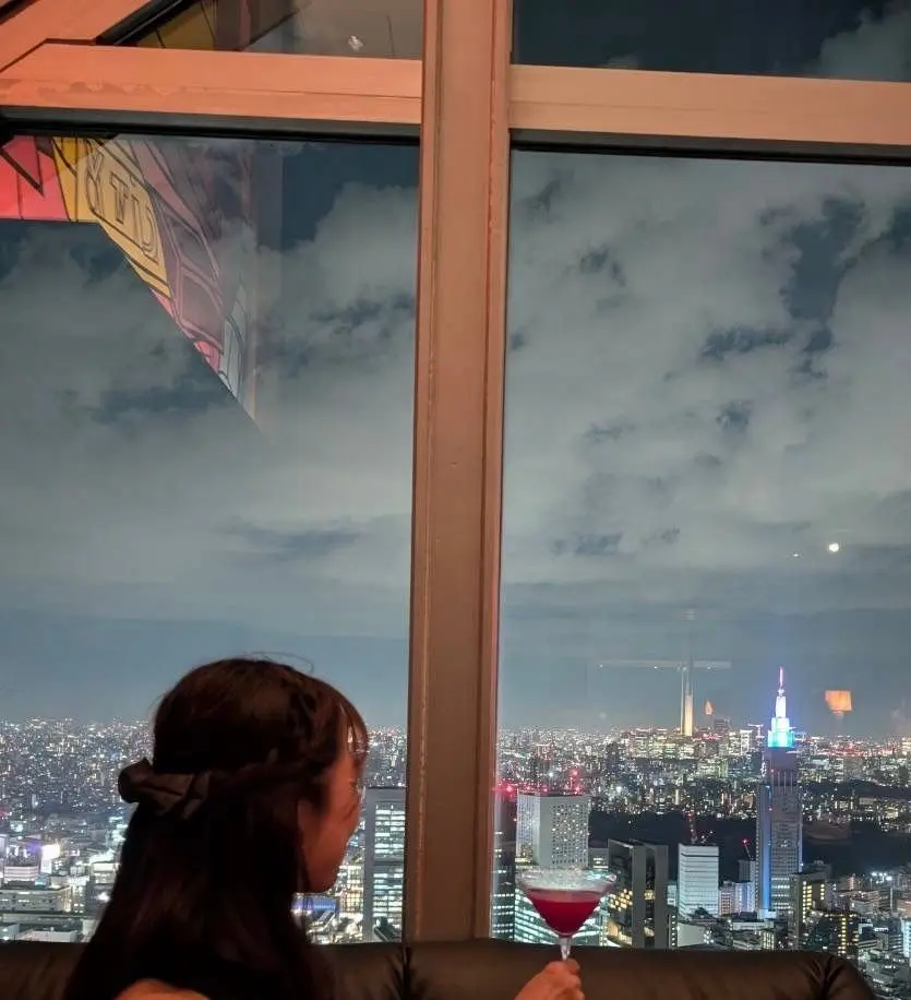 夜景を見下ろす女性。赤いカクテルを持ち、窓の外にはニューヨークの夜景と雲が広がり、遠くにエンプアイアステートビルディングが見える。