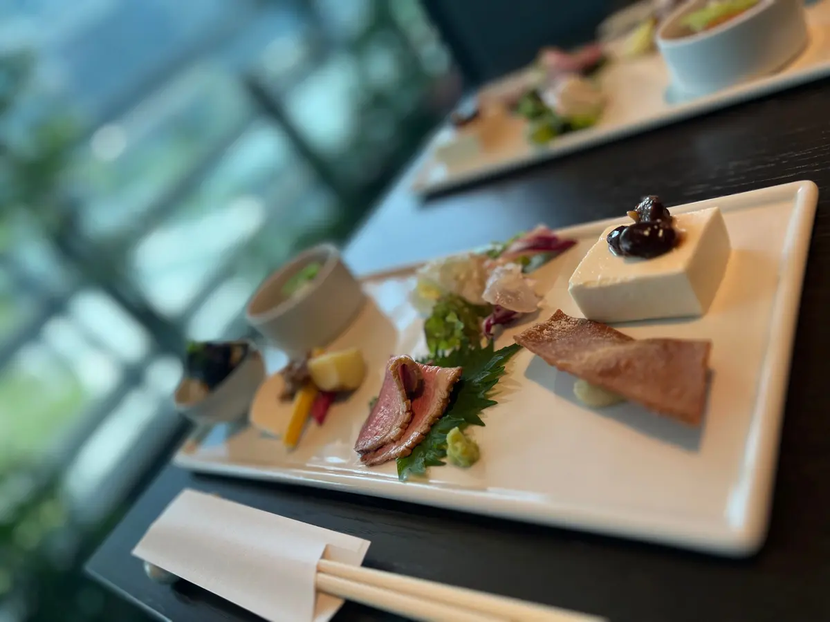 白い長方形のプレートに、肉、野菜、豆腐、焼き菓子、ソースが美しく配置された料理が並ぶ。