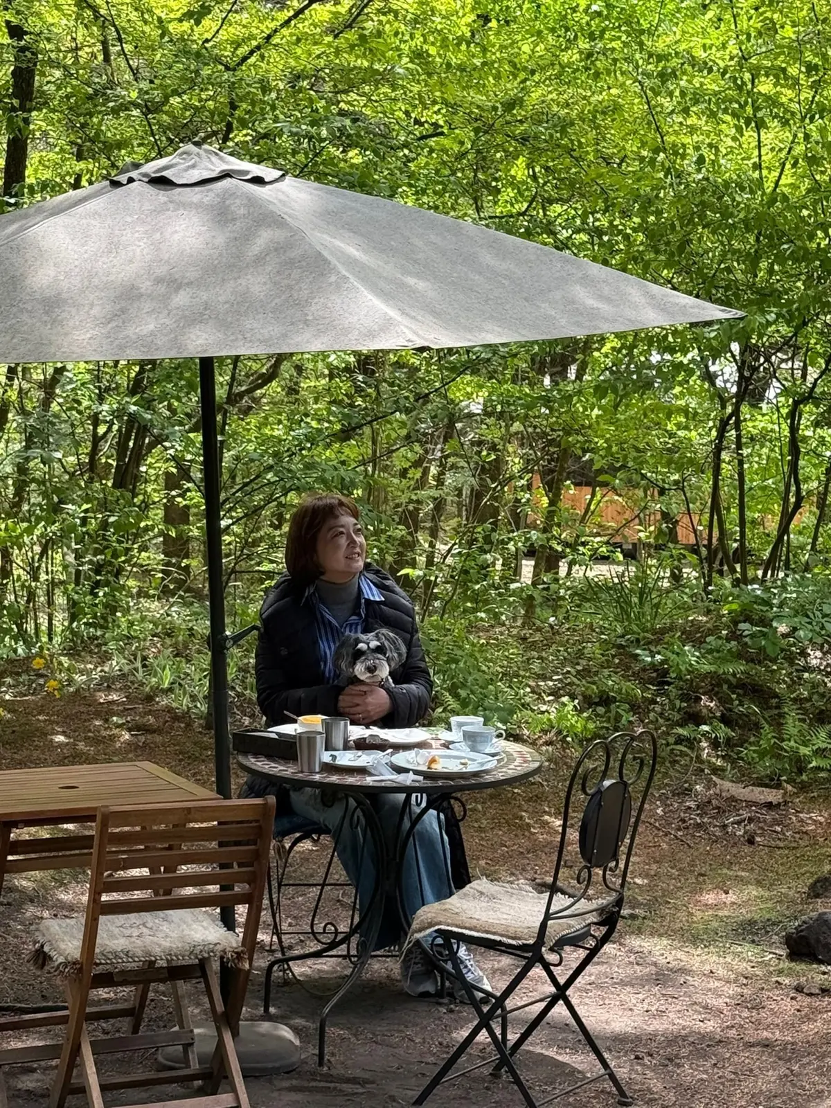 木陰のテラスで女性が犬を抱き、テーブルにコーヒーと料理が並ぶ