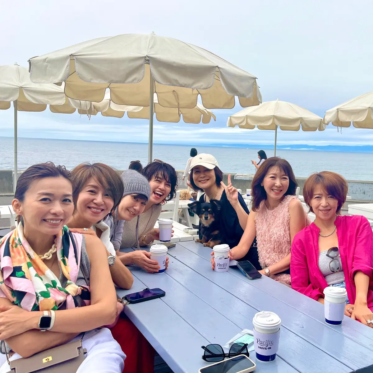 パシフィックドライブインに集合した大人女子7人。海辺のテラスで女性たちが集まり、笑顔でコーヒーカップを手にしています。テーブルには携帯電話やサングラスが置かれ、小型犬もいます。背景には海とベージュの傘が広がっています。