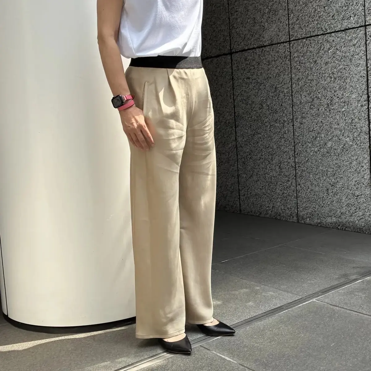 白のTシャツと黒のベルトを着用した女性が、ベージュのワイドパンツと黒いパンプスを履いて立っている全身写真。左には白い円柱状の柱、右にはグレーの壁がある。