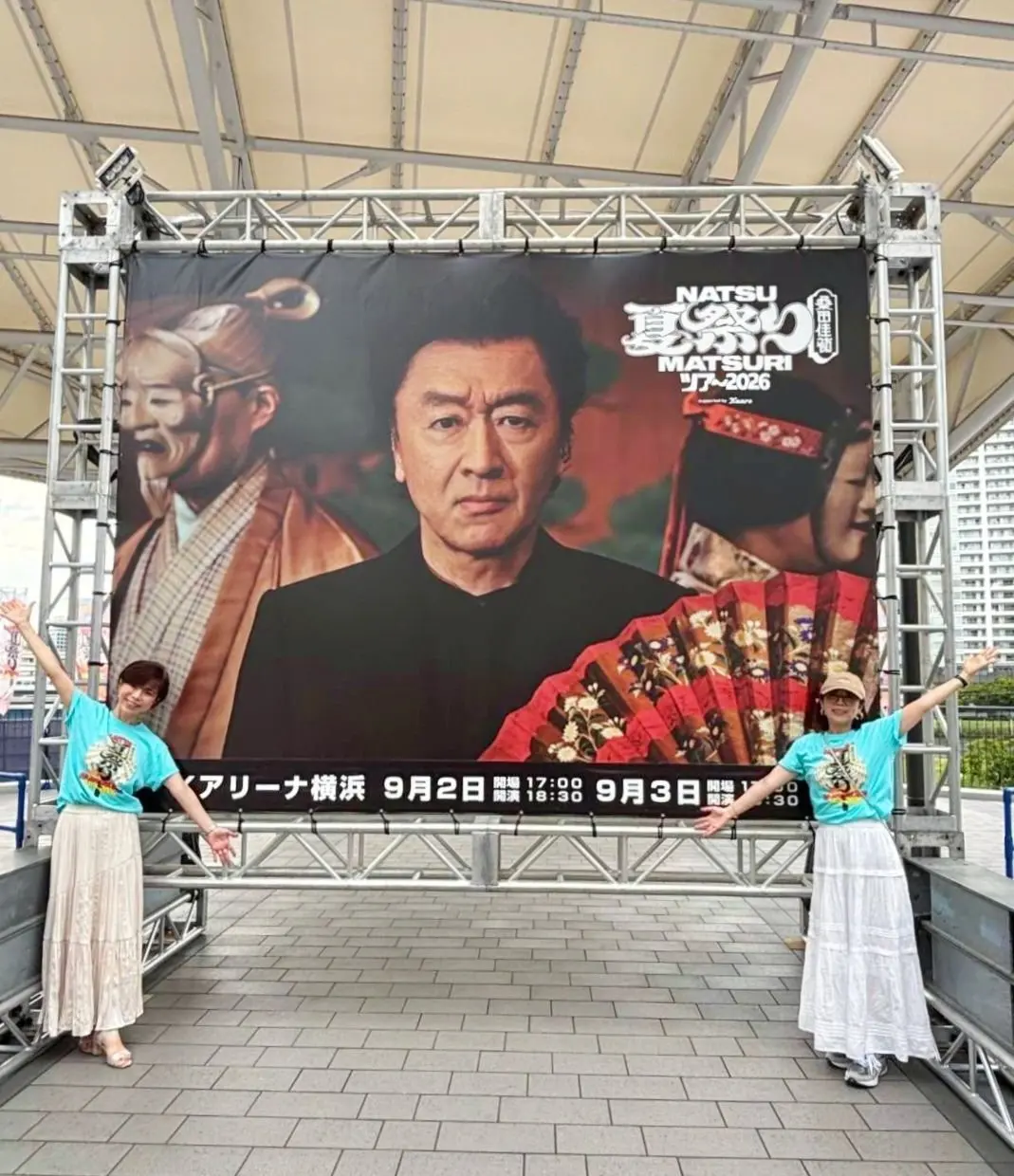 イベントのポスター前でポーズをとる2人の女性。ポスターには、男性の顔写真が大きく中央に配置され、背景に祭りの様子が写っている。「Natsu Matsuri 2026」の文字と日付の告知がある。
