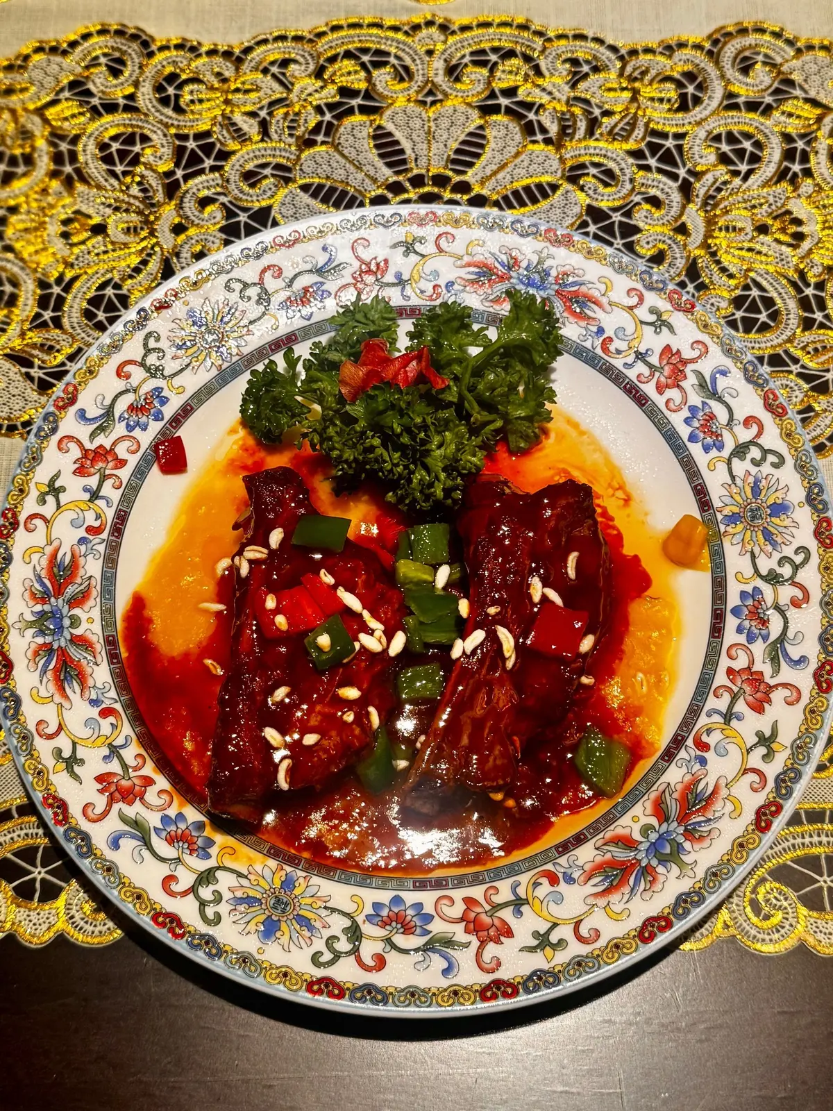 赤い色の甘辛煮込み料理が、白とカラフルな模様の皿に盛り付けられている。上には緑色の葉野菜が添えられ、白ゴマが散らばっている。