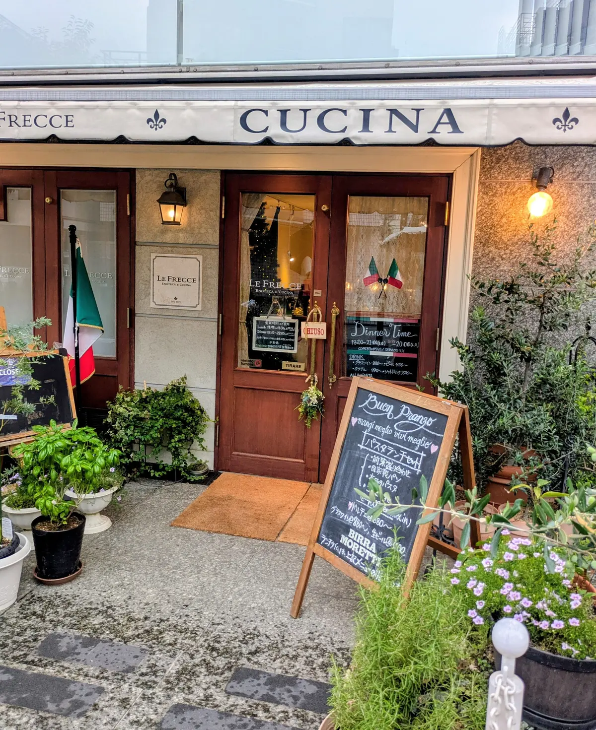 イタリアンレストラン「Le Frecce」の入り口。白と緑のストライプの看板、窓にメニューが貼られ、植木が飾られている。建物の外観はクリーム色で、雨上がりの石畳の道。