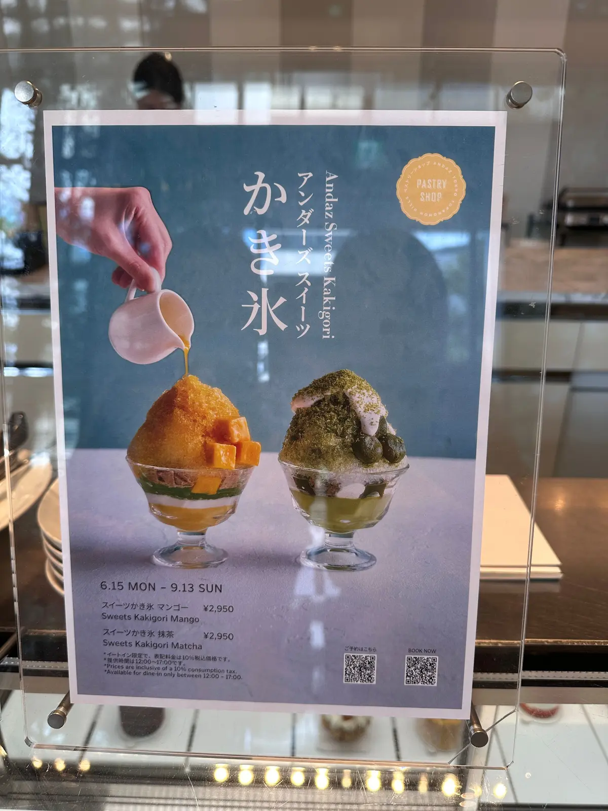 カフェの窓に設置されたメニュー。手前に「Andaz Sweets Kakigori」と書かれた案内板があり、カキ氷の写真を掲載。背景にはカフェの店内風景と外の景色が見える。カキ氷はグラスに盛り付けられ、マンゴーと抹茶のフレーバーが記載されている。