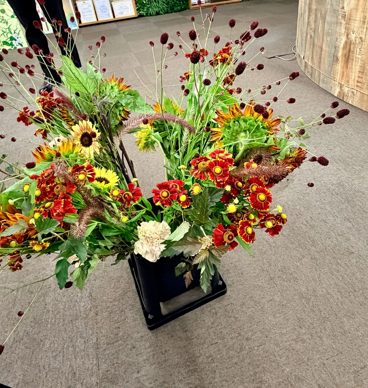 黒い立方体の花瓶に生けた、オレンジ、黄色、白の秋らしい花束。背後には緑の壁と棚があり、木製の柱が右側に立っている。
