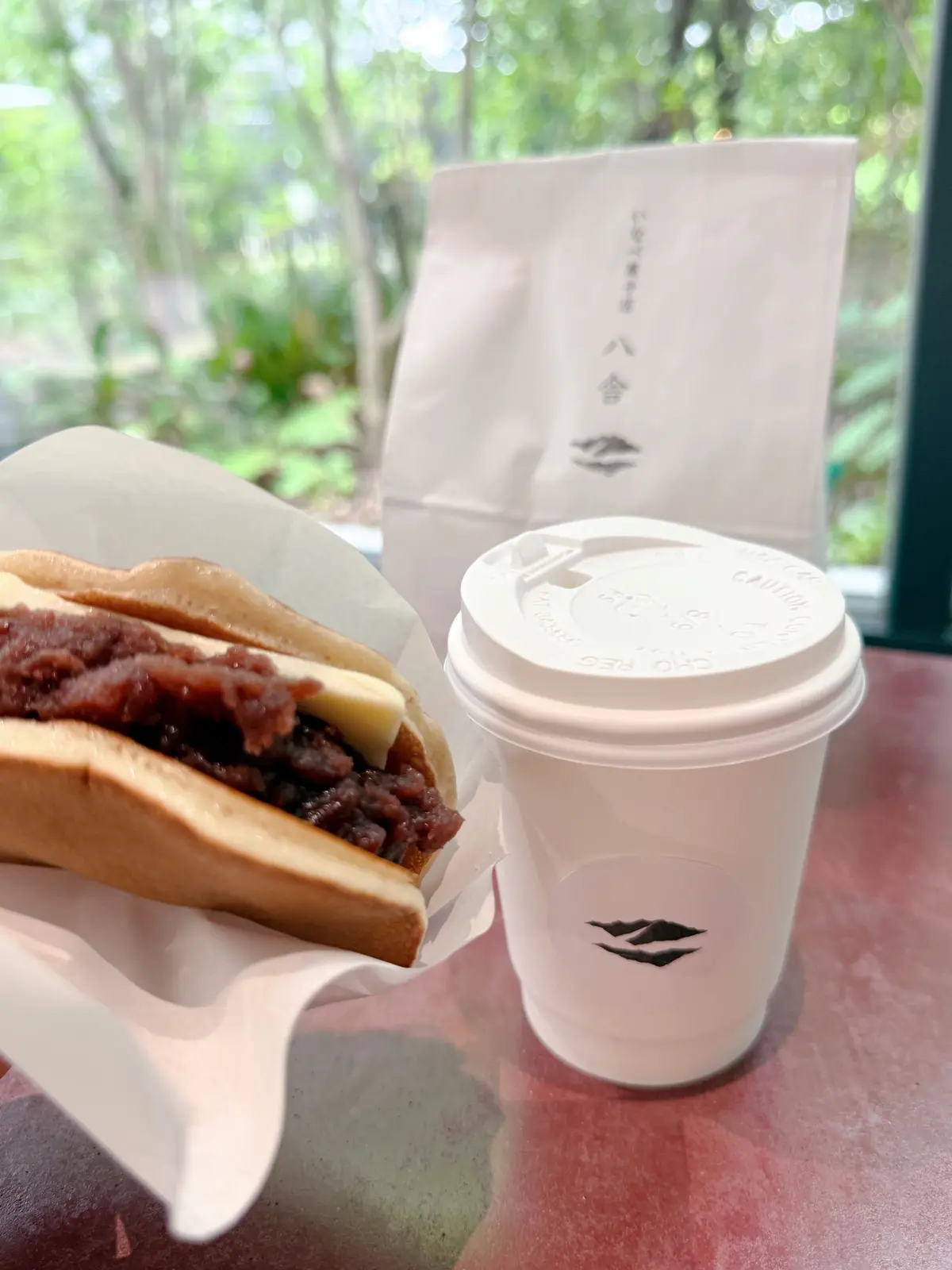 白紙に包まれたあんパンと、白いコーヒーカップが置かれている。背景には緑の植物が見える。コーヒーカップには白いロゴマークが印刷されている。