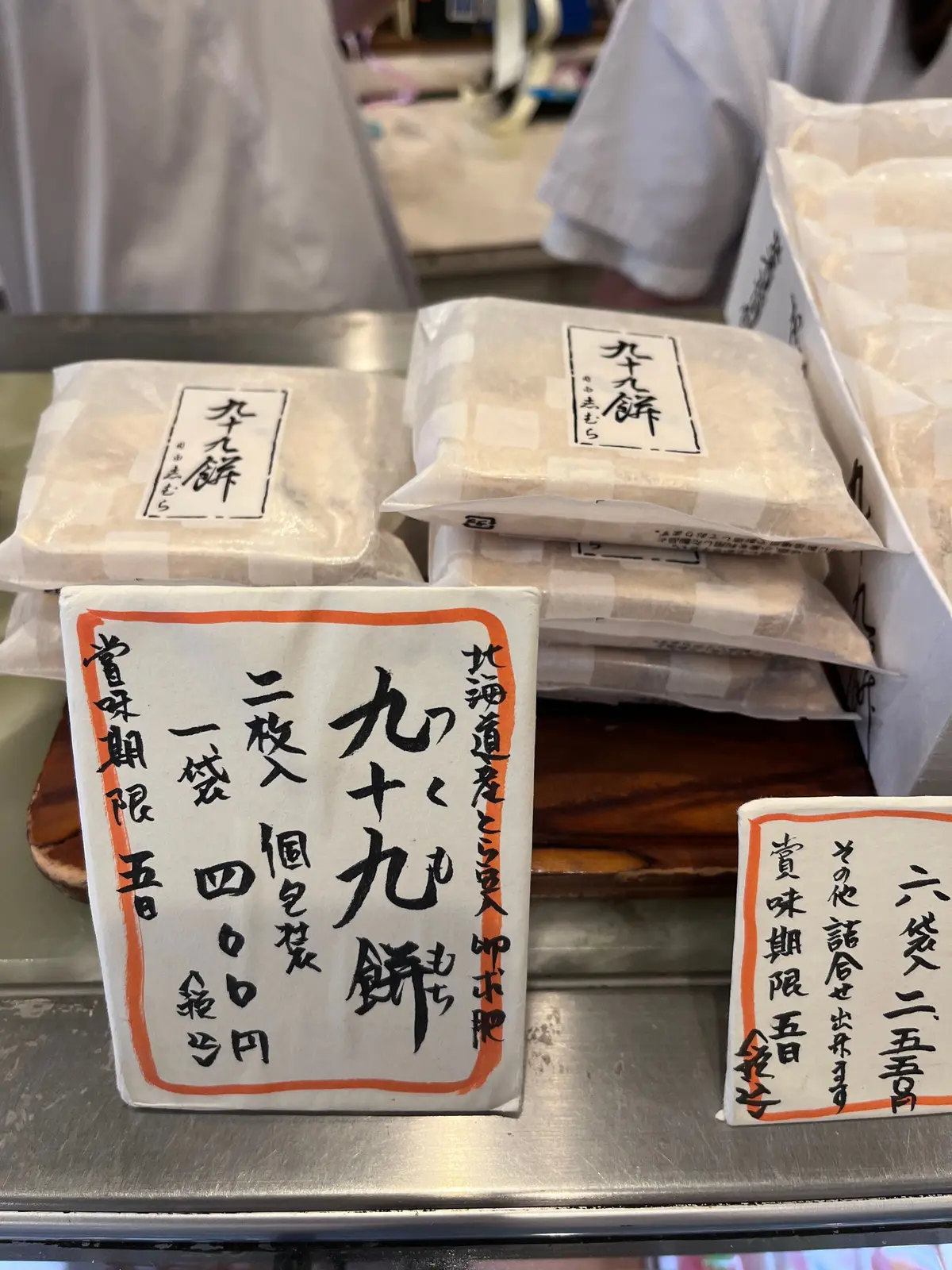 白地に黒文字の包装がされたお菓子の詰め合わせ。手前には、黄色の台紙に黒と赤の文字で価格と商品名「九十九餅」が記載されている。
