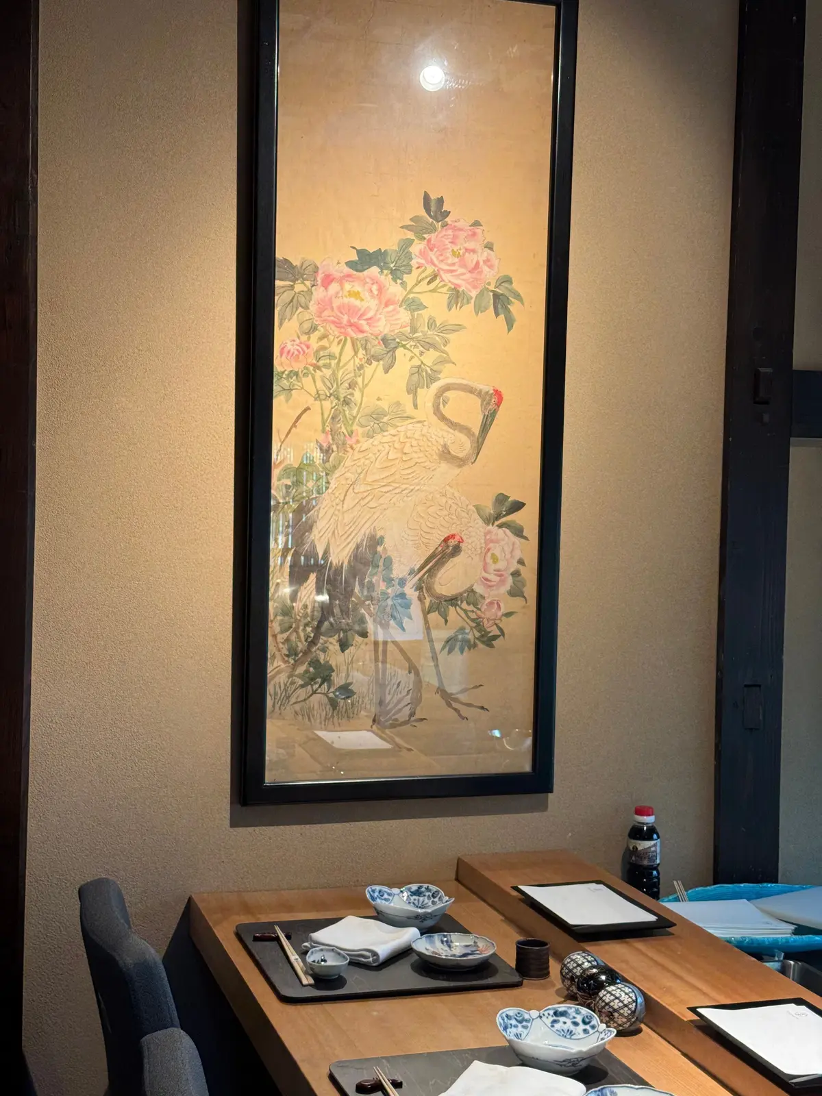 額縁に入った長方形の絵画。白鳥とピンクの牡丹の花が描かれており、背景は淡い茶色。絵の下部には岩場のような風景。テーブルには食器や調味料が置かれている。