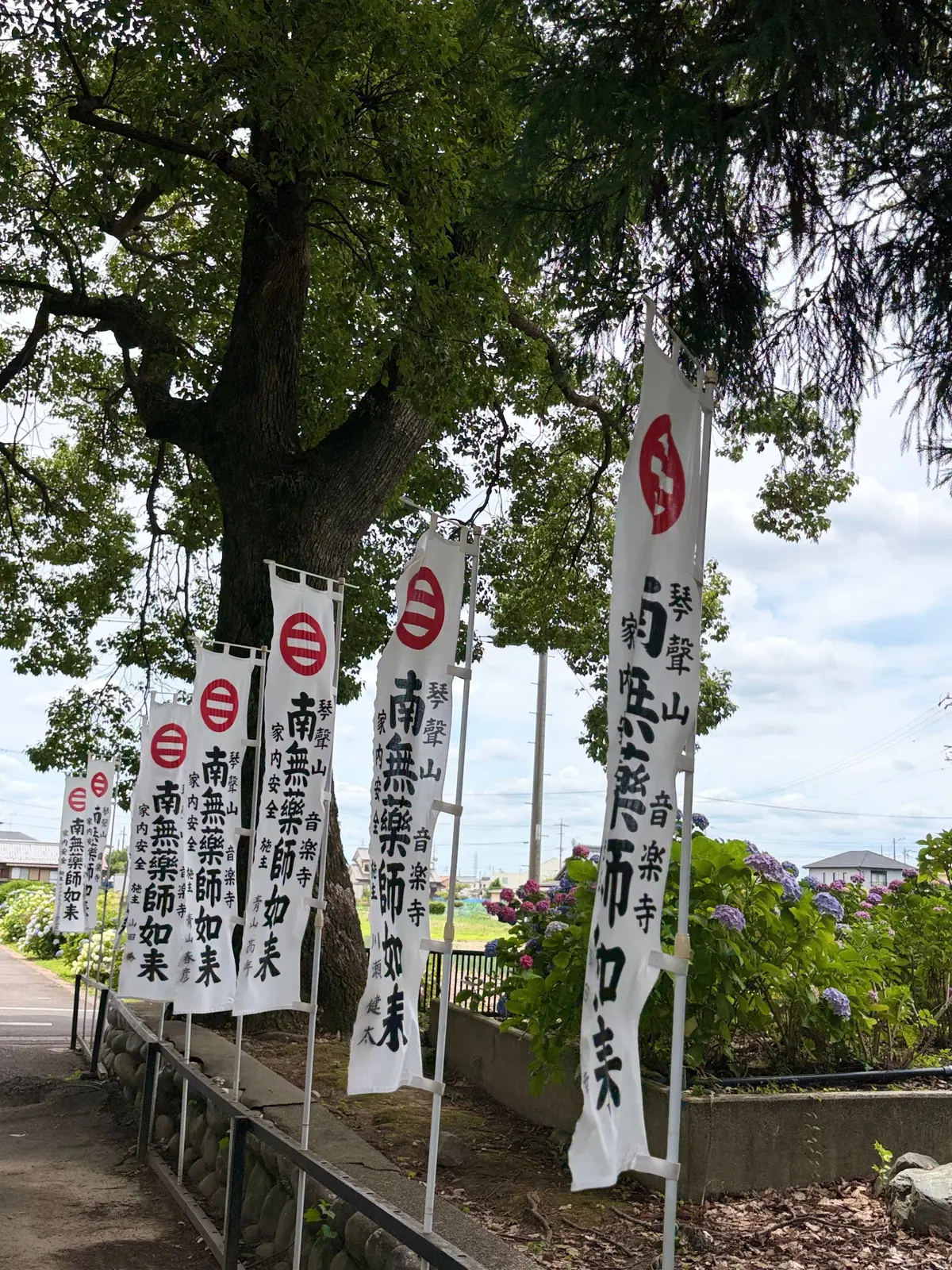 大きな木の下に、赤い印と日本語の文字が書かれた白い旗が複数立てられている。緑豊かな庭や花が周囲に広がっている。