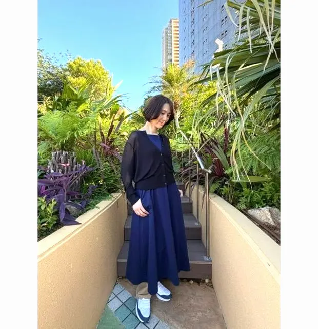 黒の長袖トップスと紺のロングスカートを着た女性が、緑豊かな植物に囲まれた階段を昇っている。背景には高層ビルが映る。