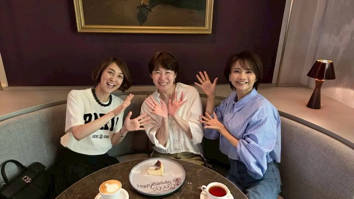 3人の女性がテーブルに座り、笑顔で手を広げています。テーブルの上にコーヒーとケーキがあり、ケーキには「Happy Birthday」の文字が書かれています。背景には絵画とランプが置かれています。
