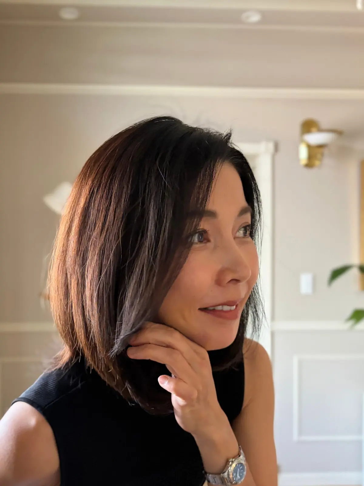 黒いネックラインのトップスを着た女性が、横を向いて微笑んでいる。茶色のボブヘアに、銀色の腕時計をしている。背景は白い壁で、金色の壁掛けライトが見える。