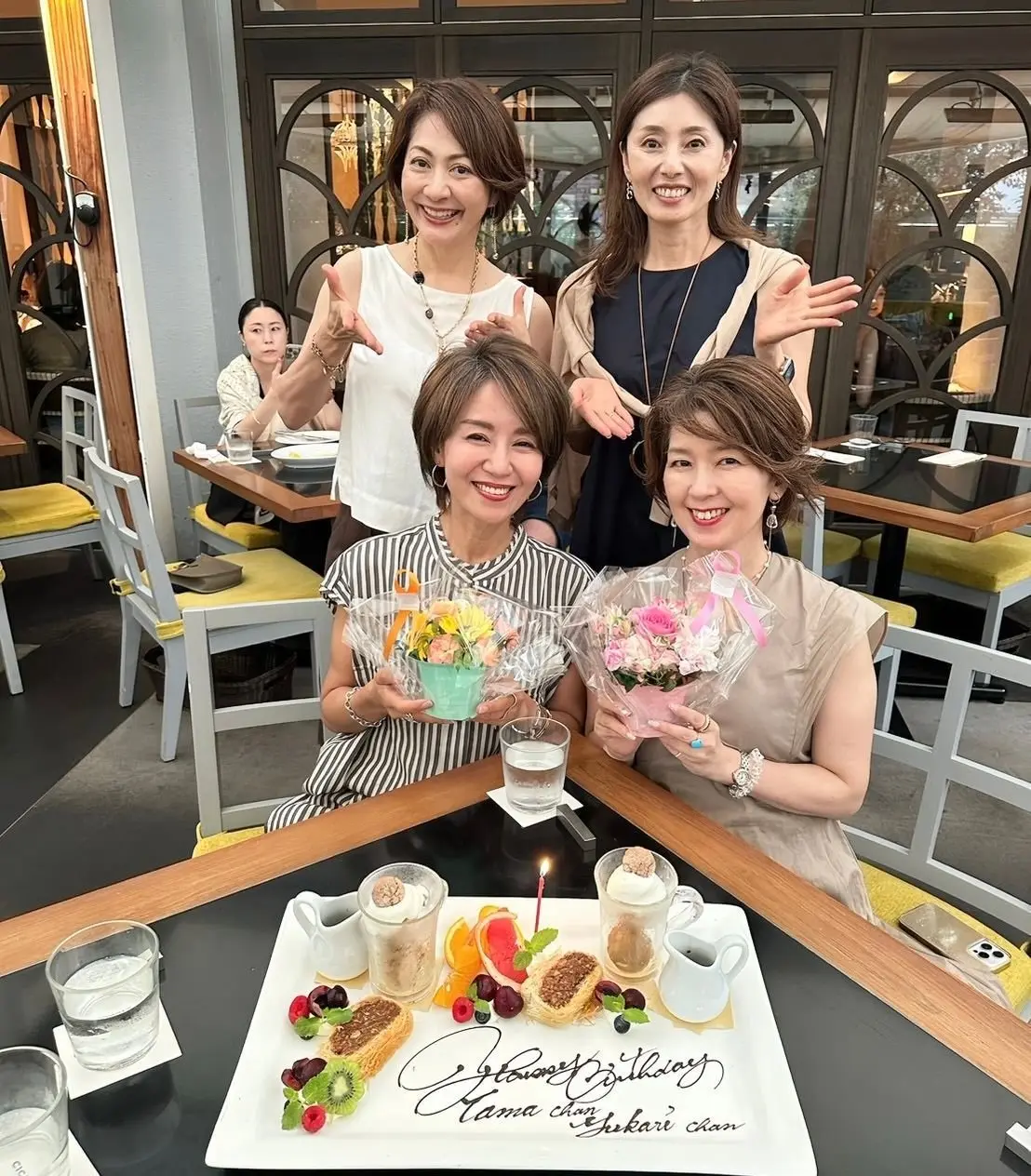 レストランで誕生日のお祝いをする4人の女性。手前に座る2人は花束を持ち、中央にはフルーツや飲み物が盛り付けられたプレート。背景には他の客とスタッフが見える。