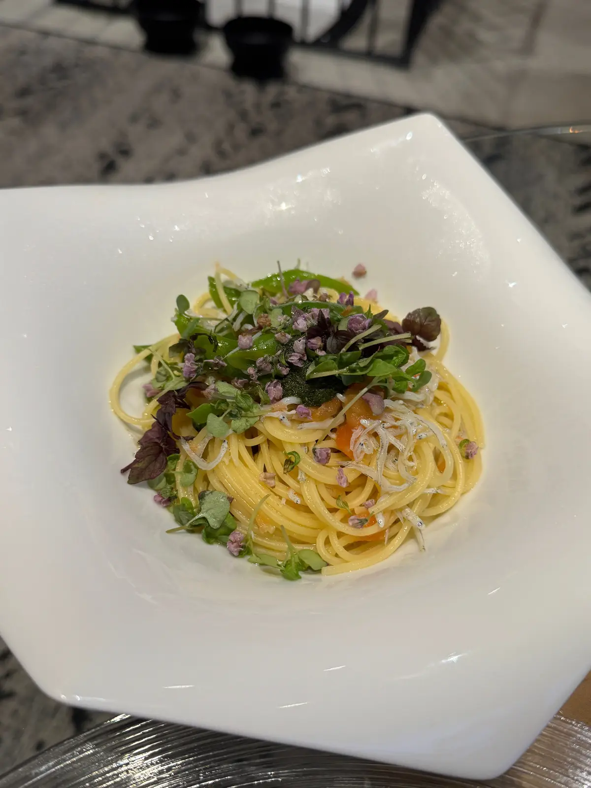 白磁の皿に盛り付けられた、黄色のパスタ。表面には緑や紫のマイクロハーブ、白っぽい魚介類が散らばっている。