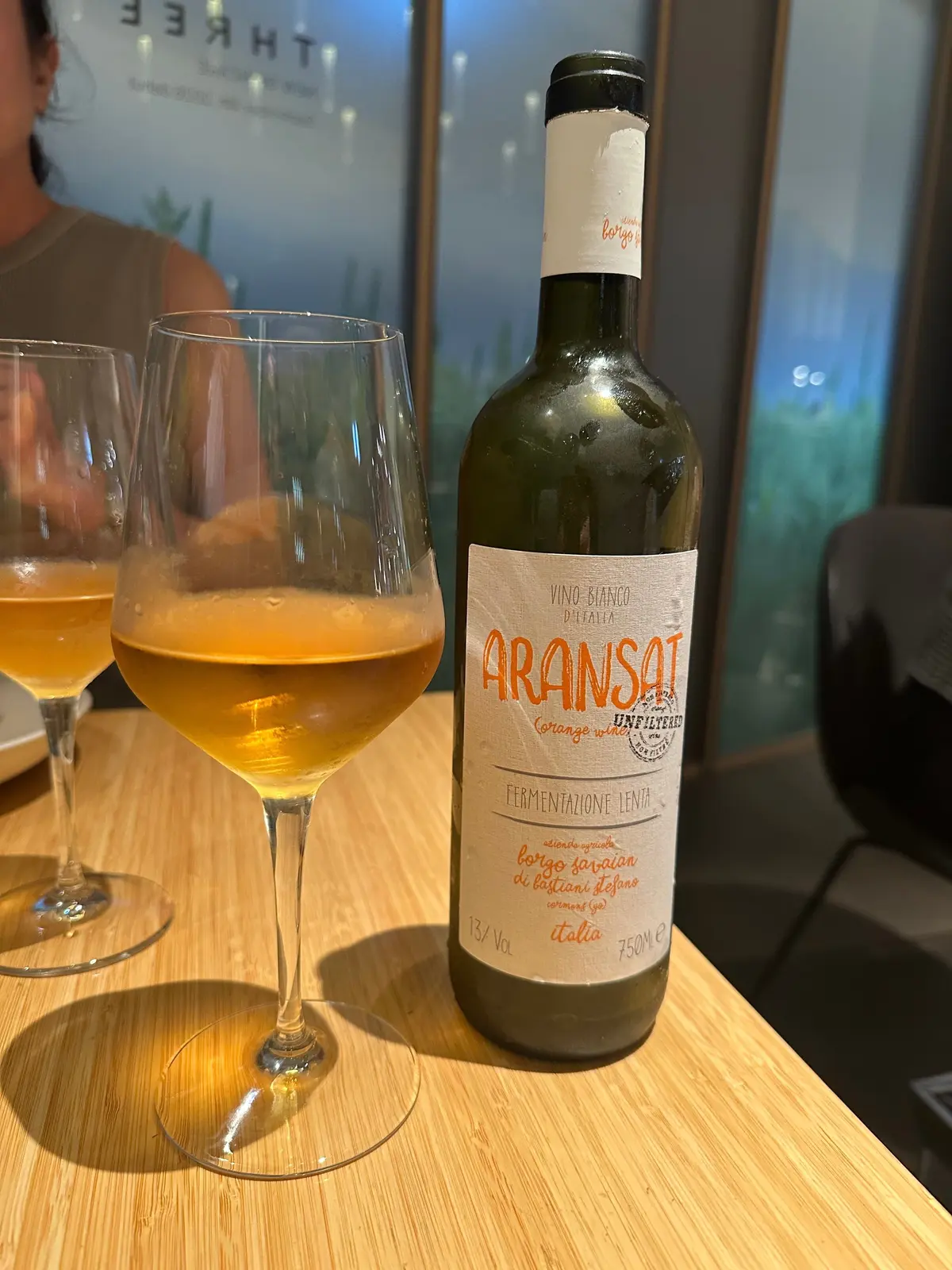 木製のテーブルに置かれた、オレンジ色のワインとグラス。ワインボトルには「ARANSAT」のロゴとイタリア語のラベルが貼られている。グラスには琥珀色のワインが注がれており、光を受けて輝いている。