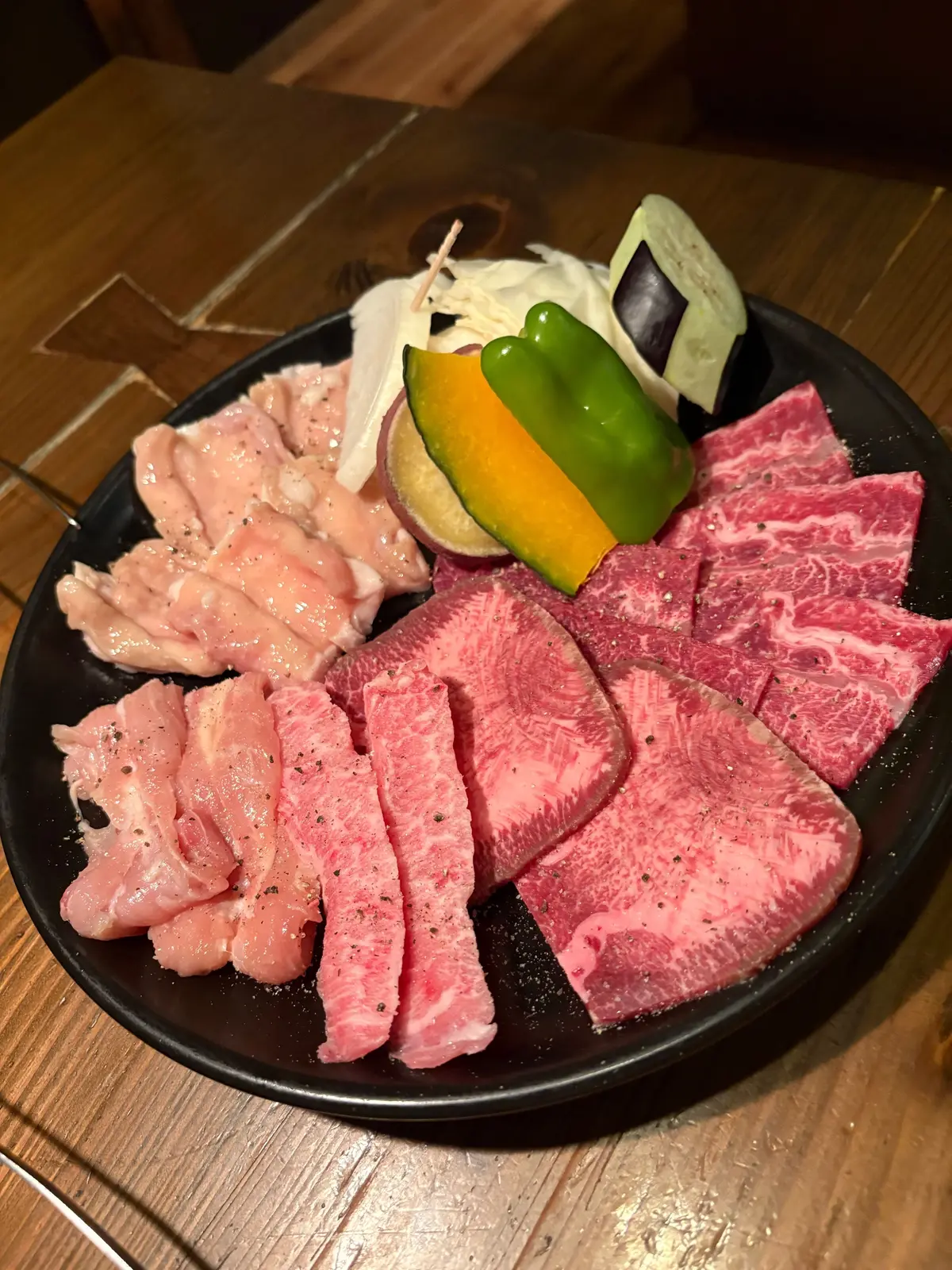 黒い皿に生肉と野菜が並ぶ。赤身の肉が中心で、緑と黄色のピーマン、なす、玉ねぎが添えられている。