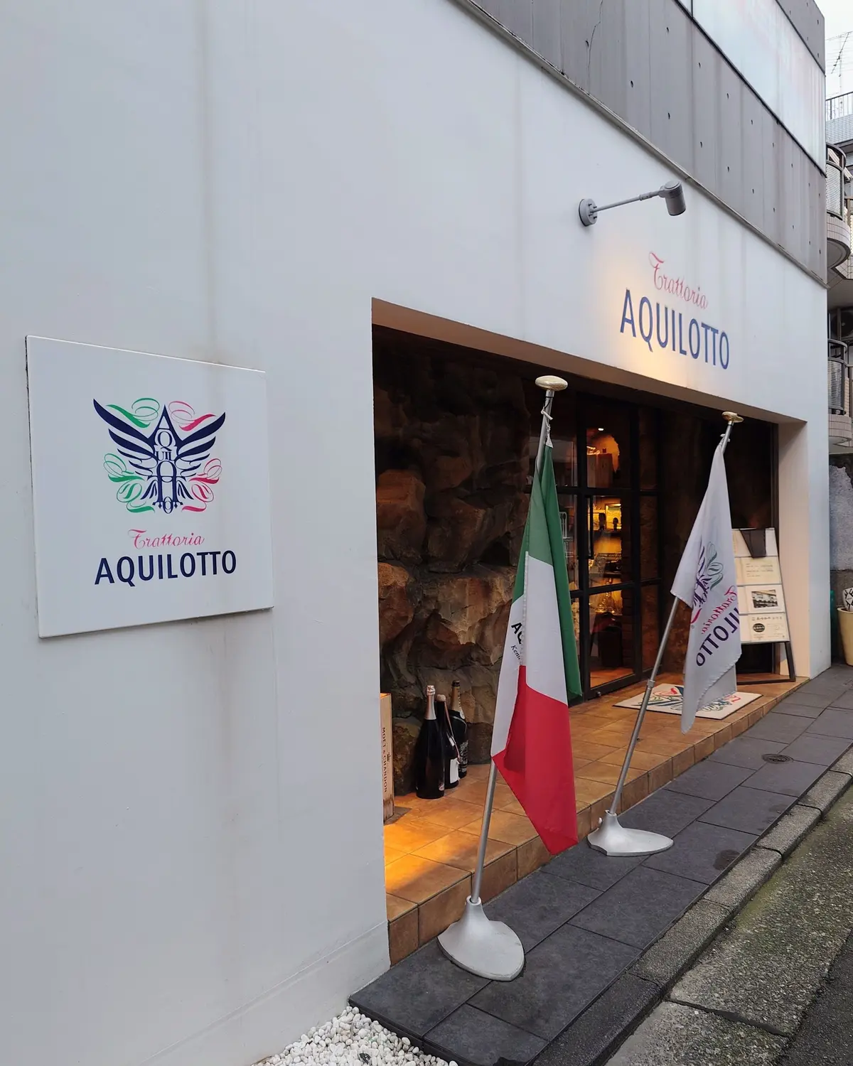 白い壁に「トラットリア アクイロット」のロゴと看板があるイタリア料理店。入り口にはイタリア国旗とお店の旗が立てられ、ワインボトルが置かれている。