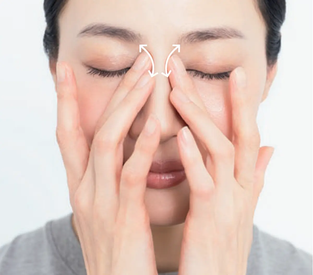 老廃物がたまりやすい 鼻根横はかき出すように 両手で目の周りをさすり、目の疲れを和らげる女性。白い背景に、目の上部をさする動きが矢印で示されている。