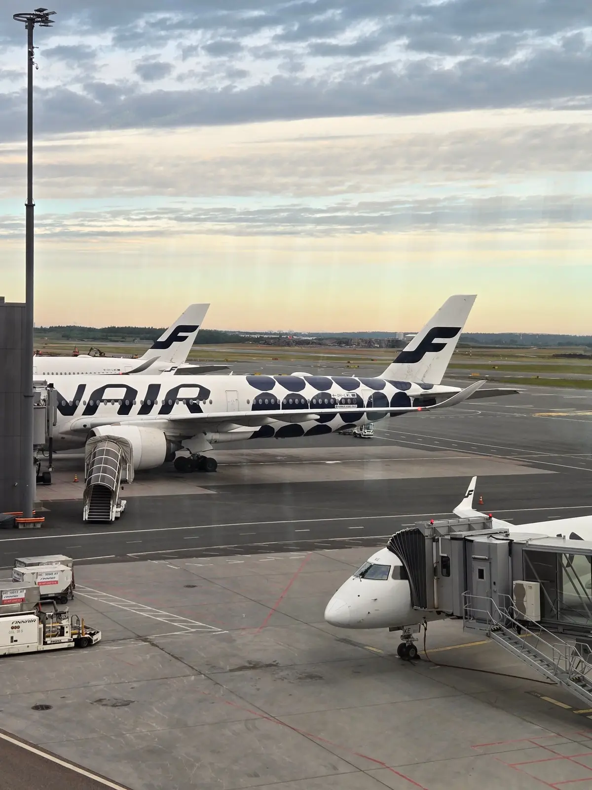 Wizz Airの白と青の旅客機が駐機している空港の風景。機尾にはWizz Airのロゴマークが描かれ、前方のジェット機と搭乗ブリッジが見える。背景には曇り空と遠くの緑地帯が広がる。
