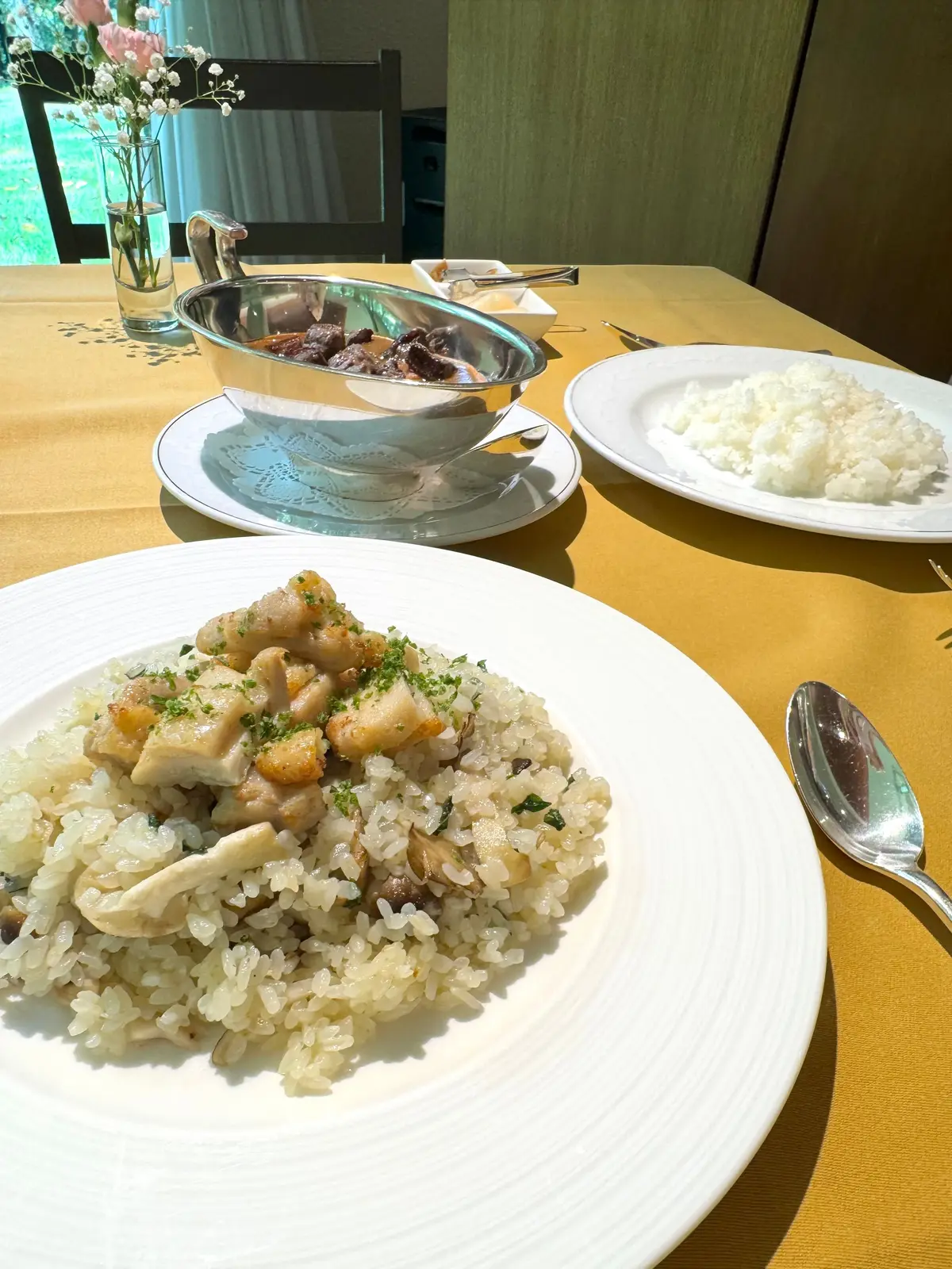 黄色のテーブルに置かれた和食の盛り合わせ。手前には鶏肉とキノコが乗ったご飯、奥には白米と煮物、小さな器に入った薬味、花瓶に入った花が置かれている。
