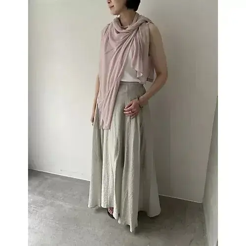 ピンクのラップ風カーディガンにグレーのワイドパンツを着た女性が、壁に寄りかかり、右手を腰に置いている。