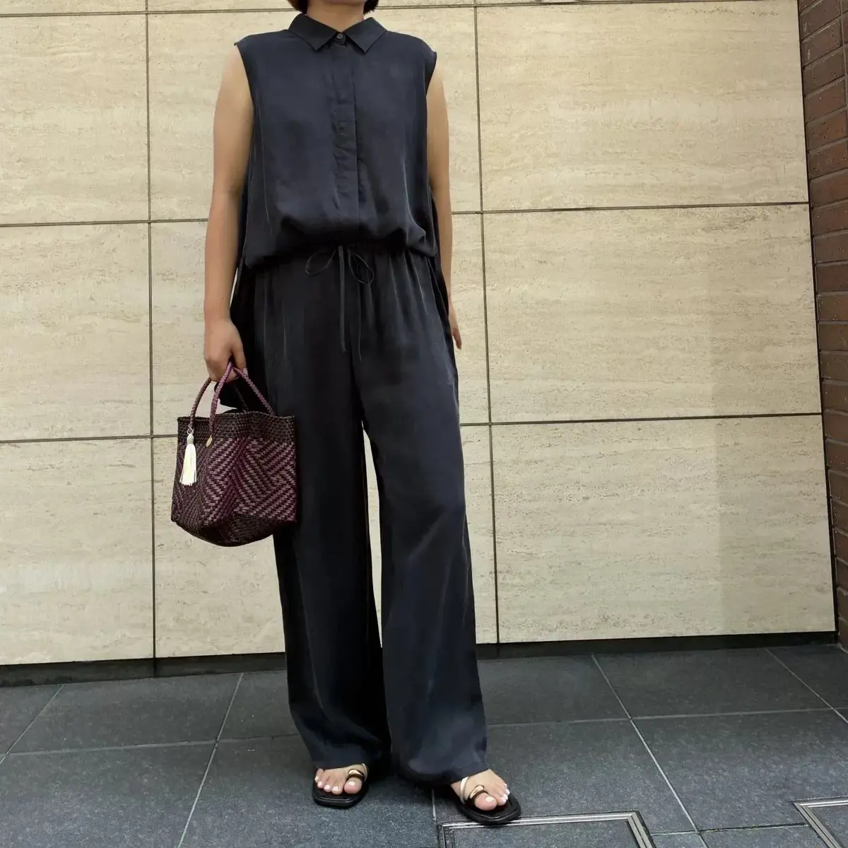 黒色のスリーブレスシャツとワイドパンツのセットアップを着用した女性。茶色の編み込みバッグを持ち、黒いサンダルを履いている。背景はベージュ色のタイル壁。
