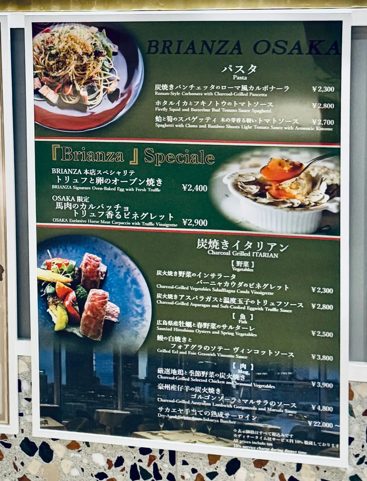 大阪のレストラン「BRIANZA OSAKA」の入り口。右側にメニューが額装された緑色の装飾パネルがあり、左上にはブランド名が光るサイン、上部には矢印の標識がある。壁はそれぞれ白い大理石調と茶色のレンガ調。