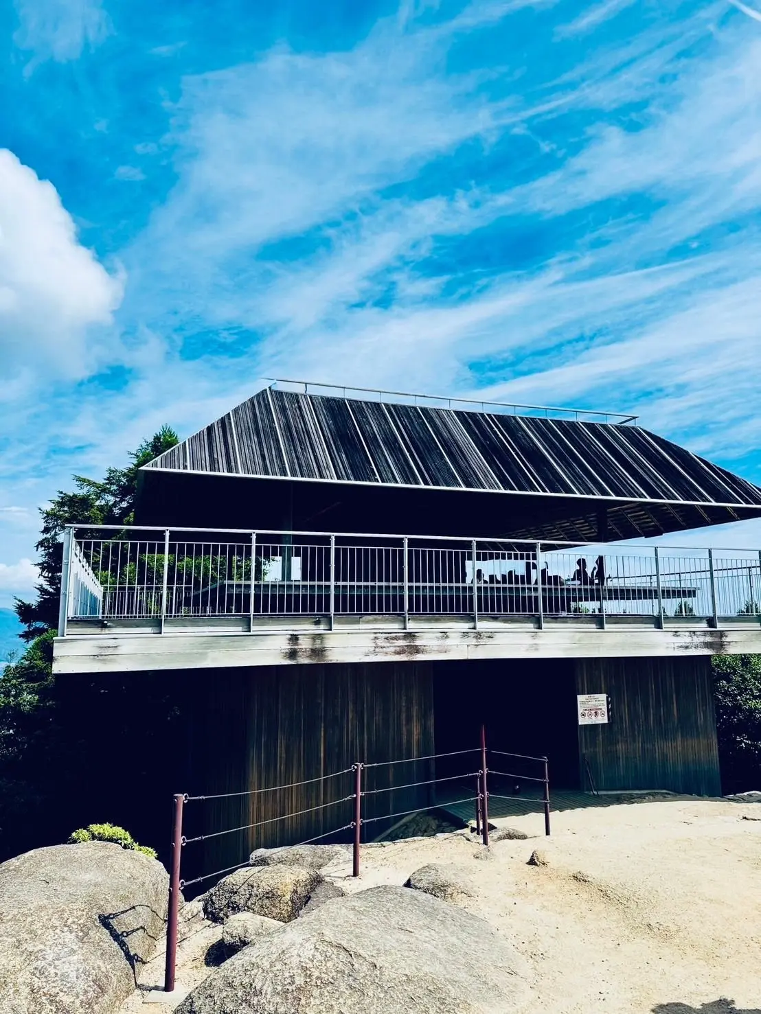 木造の休憩所が、砂利と岩の上に建っている。屋根は濃いグレーで、手すりが付いたテラスがある。背景には青空と白い雲が広がり、緑の木々が見える。