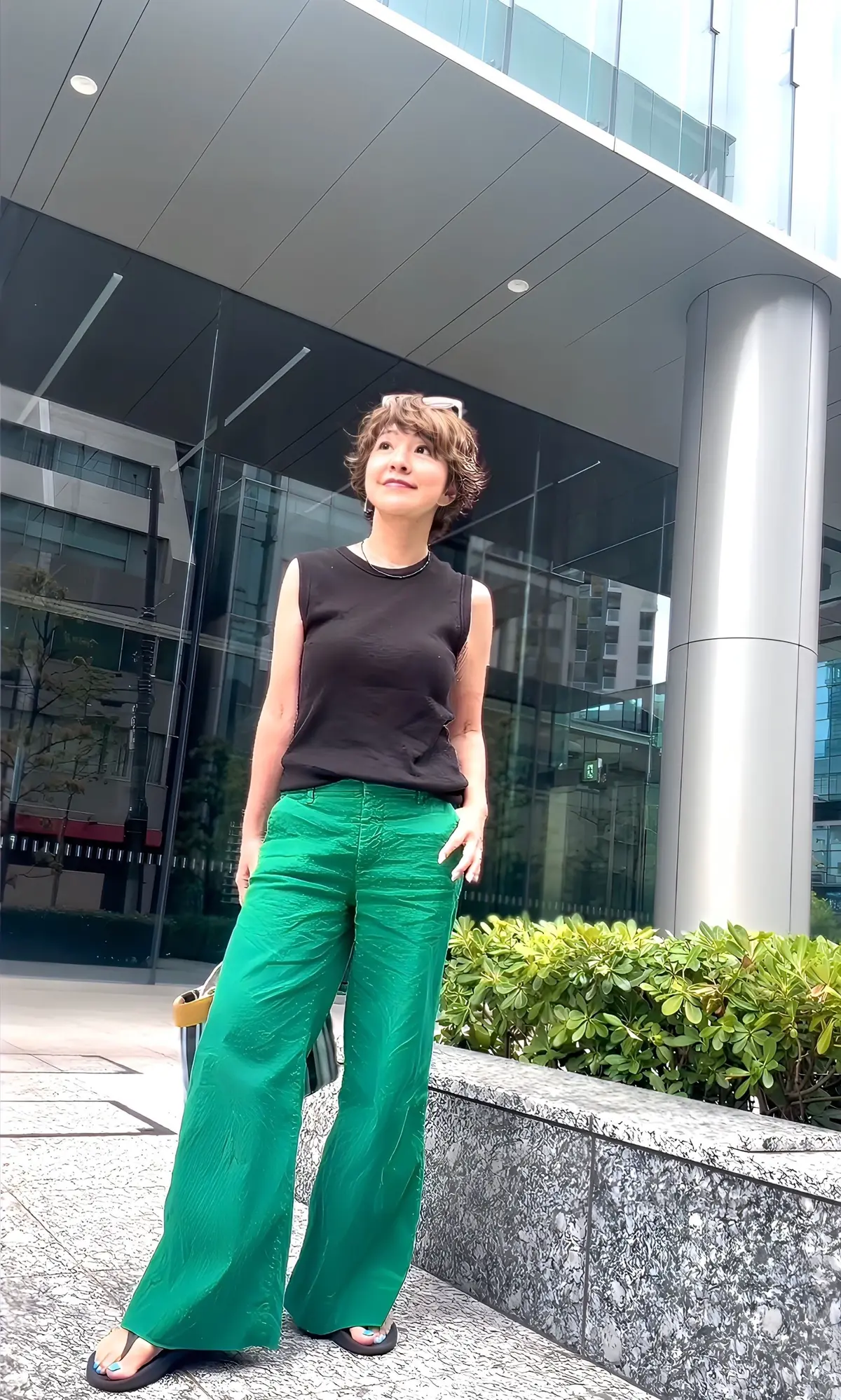 黒いタンクトップにエメラルドグリーンのワイドパンツを着用した女性。茶色の短い髪型で、建物の前でポーズをとっている。足元はサンダル。