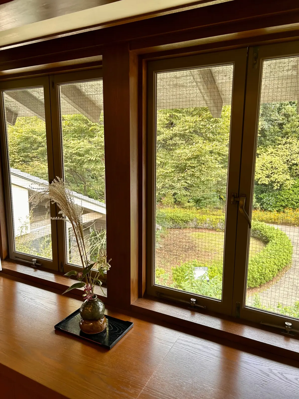 木製の窓枠に設置された、緑色の網戸付きの窓が二つ並んでいる。窓の外には緑豊かな庭園が見える。窓の台には、黒い台座の上に小さな花瓶に入った植物が置かれている。