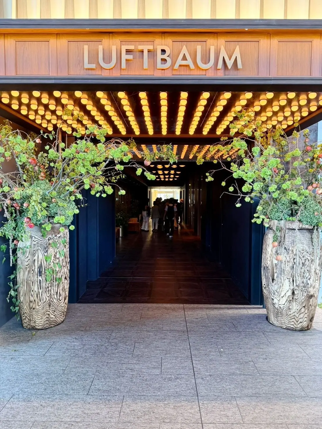 「Luftbaum」と書かれた看板の下、光の装飾が輝く入り口。左右には木製の植木鉢に緑の植物が植えられている。通路は奥へと続いており、人々が歩いている。