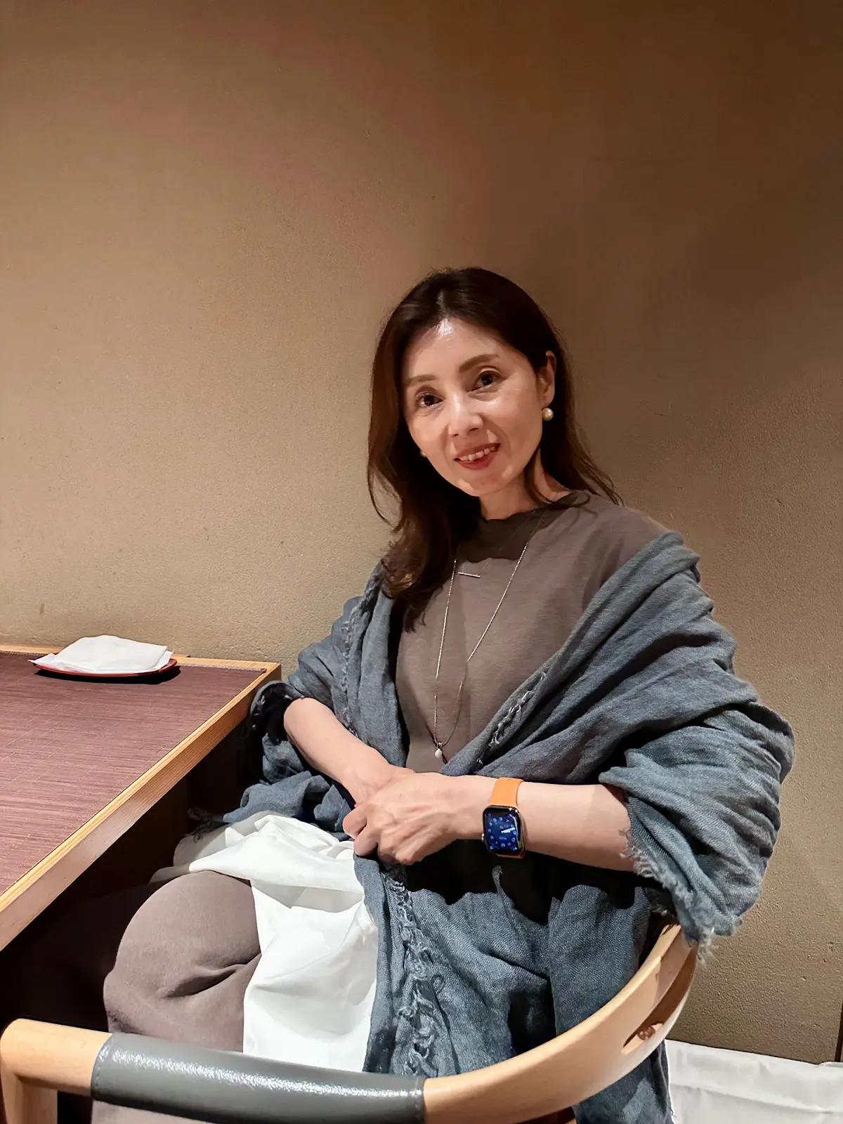 茶色い壁の背景に、木製の椅子に座る女性。グレーのデニムジャケットを羽織り、茶色のトップス、スマートウォッチを着用。右手には白い布が置かれ、左手には白いナプキンがあるテーブルが隣接。