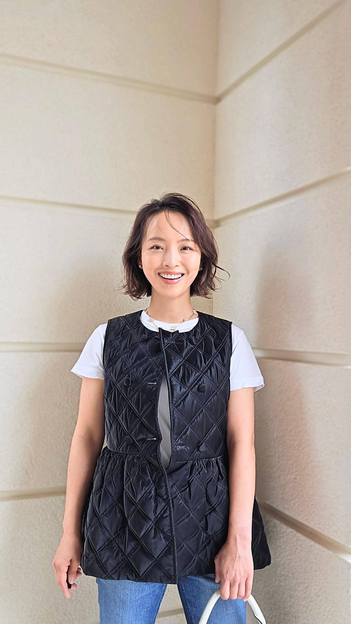笑顔の女性が、白いTシャツに黒いキルティングベスト、青いジーンズを着用して立っている。茶色のボブヘアで、背景はベージュ色の壁。