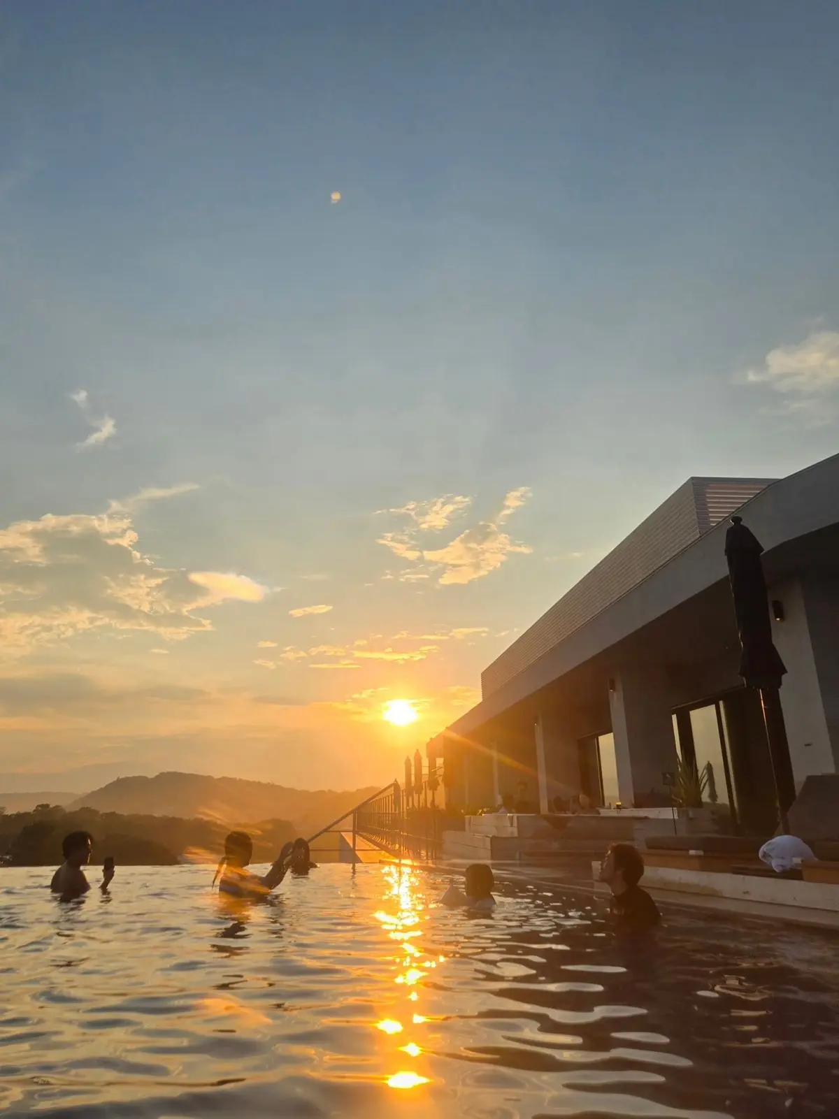夕焼け空の下、プールで遊ぶ人々。水平線に沈む太陽が水面に反射し、オレンジ色の光が広がる。背景には建物と緑豊かな山々が見える。