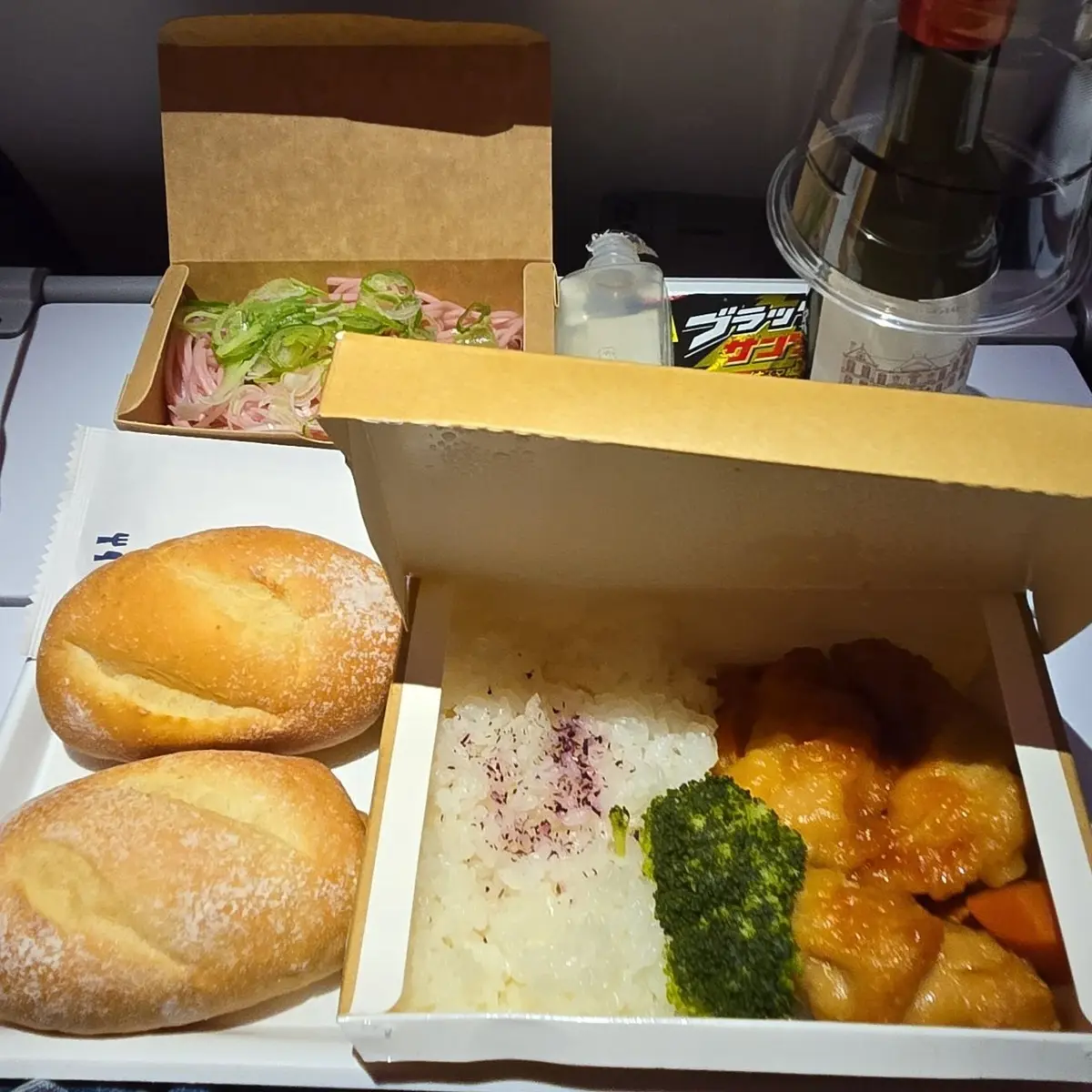 航空機内での食事。白の弁当箱に白米、鶏肉の照り焼き、ブロッコリー、パン、付け合わせの和え物、飲み物、おしぼりなどがセットされている。